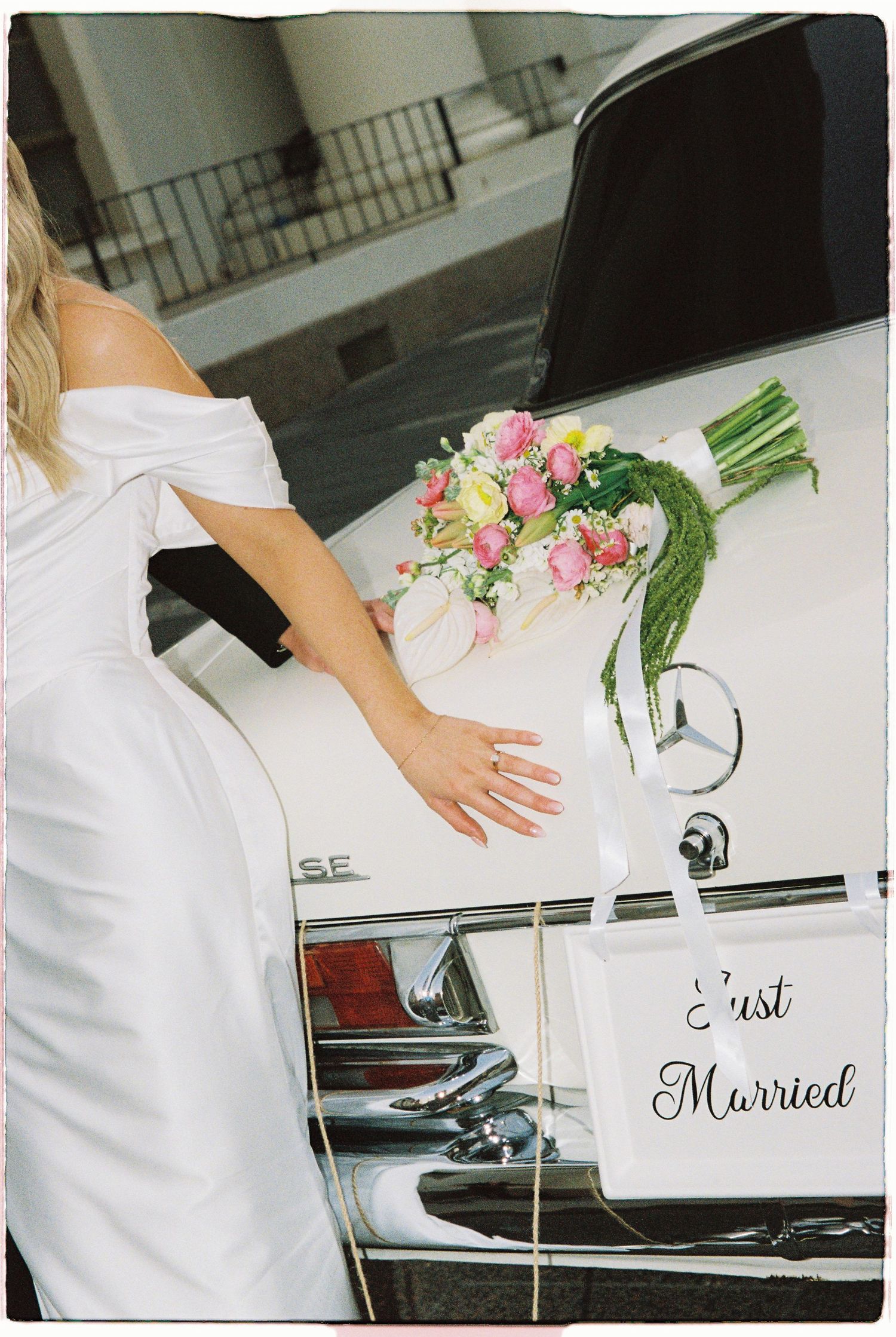Bride in white dress placing bouquet on vintage white Mercedes Just Married wedding car.