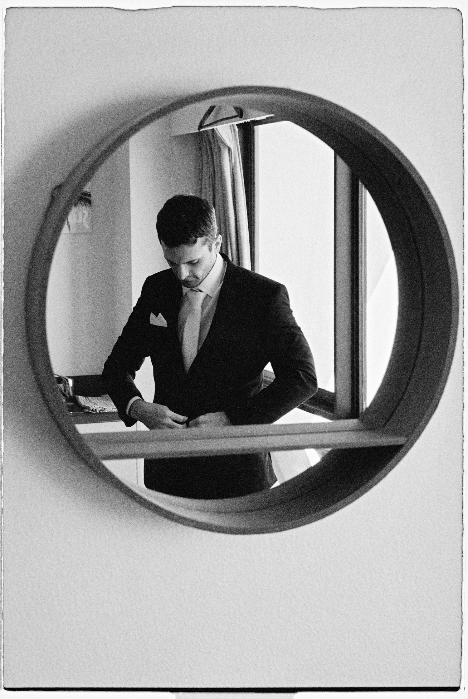 Groom in black suit adjusting tie reflected in a round circular mirror, black and white photo.