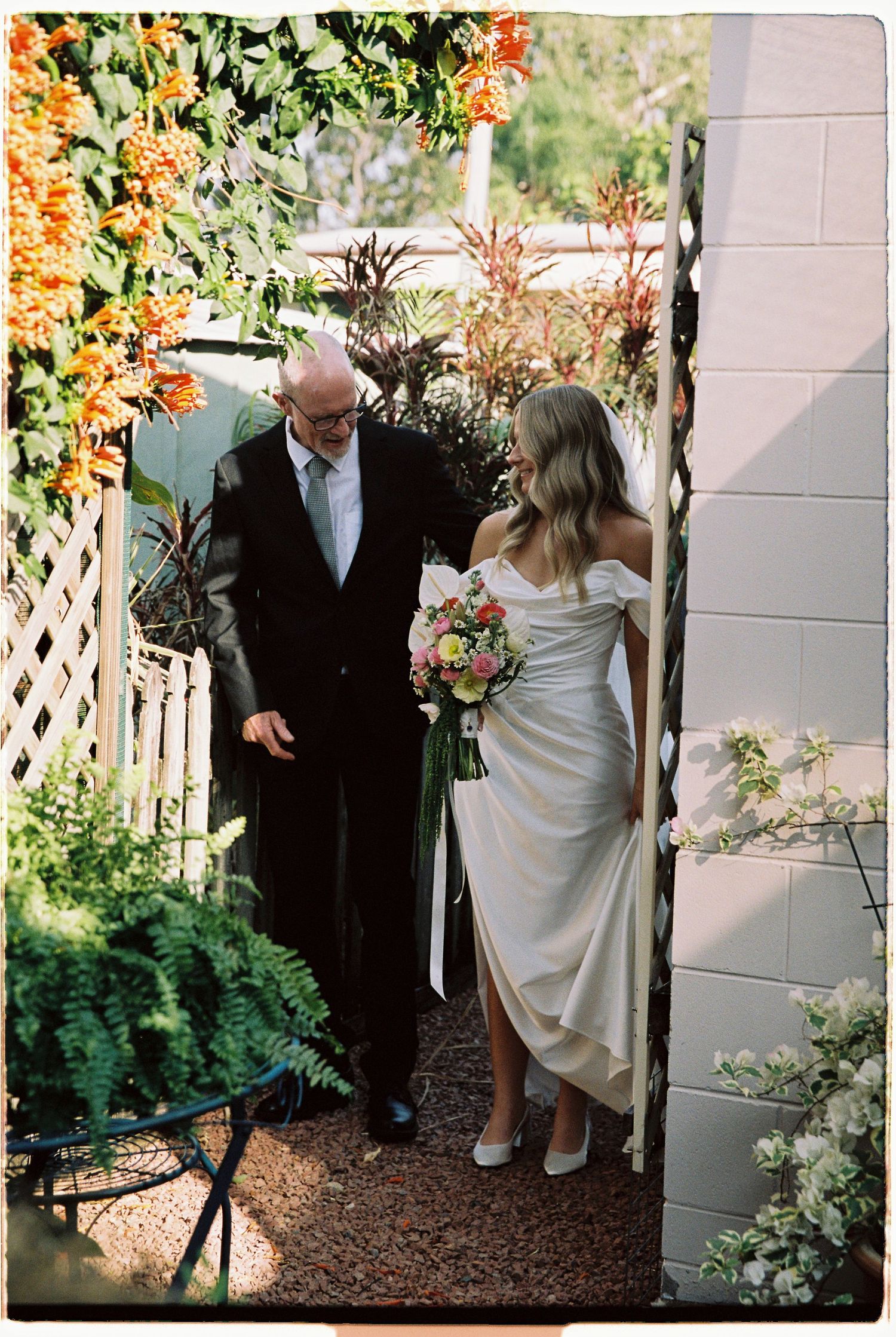 Bride in white off-shoulder gown walking with older gentleman through a flower-lined garden path.
