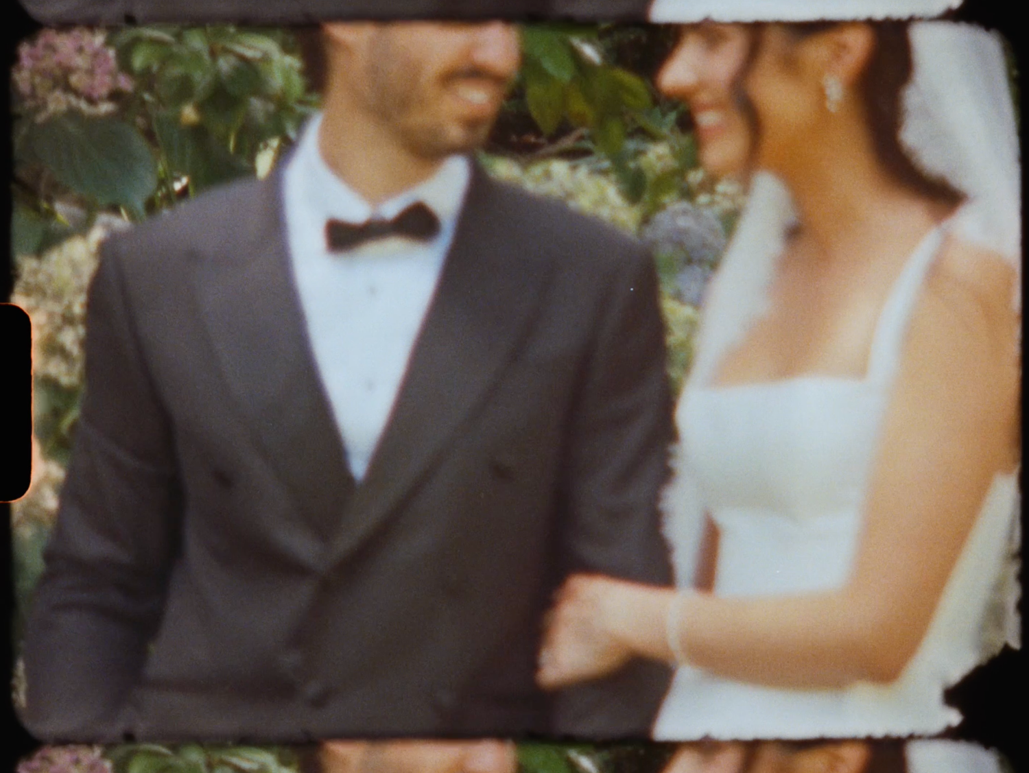 Smiling bride and groom in a dark suit with bow tie posing together in a lush garden setting.