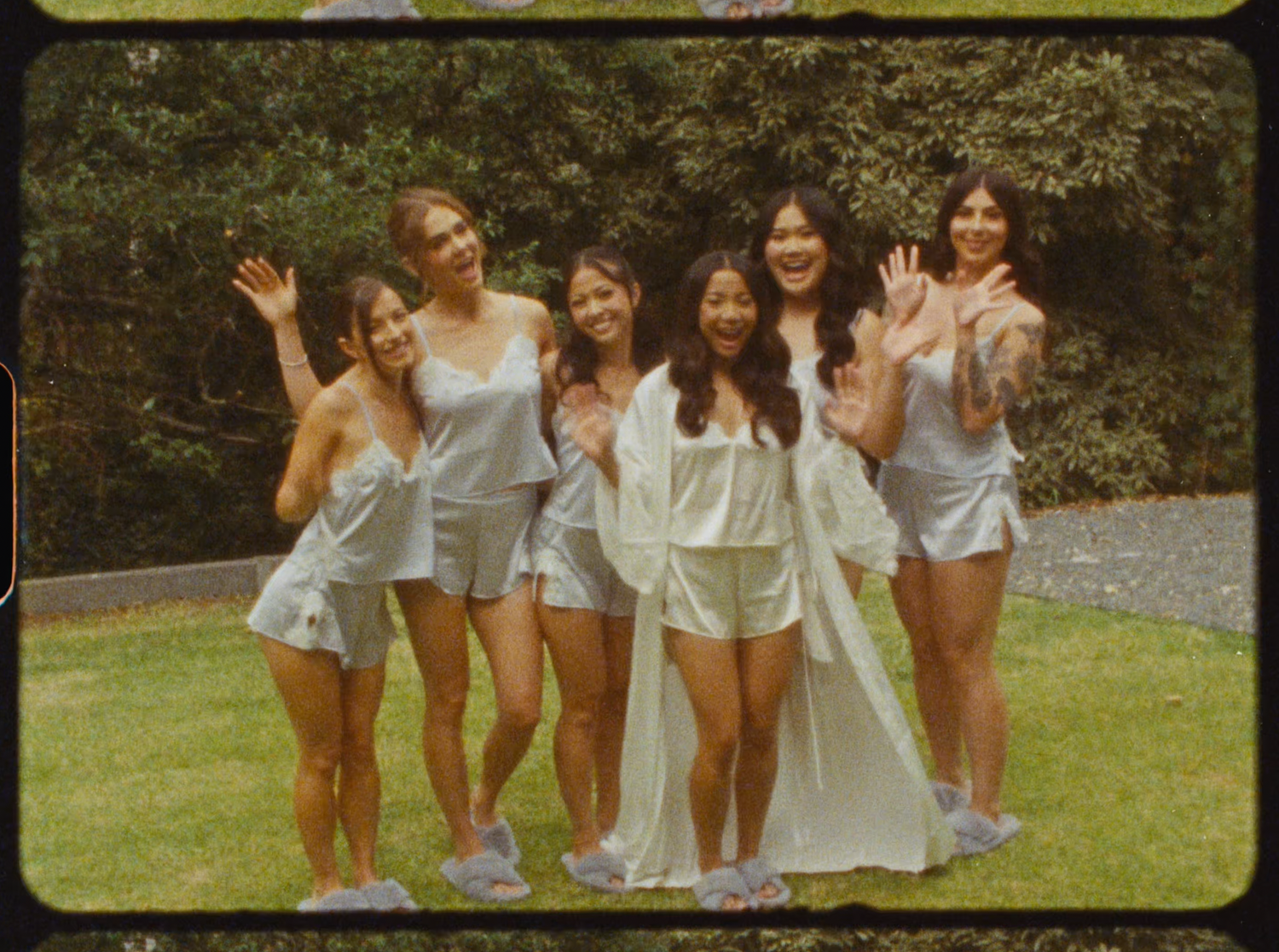 Six bridesmaids in matching blue satin sets waving and posing on a sunny green lawn.