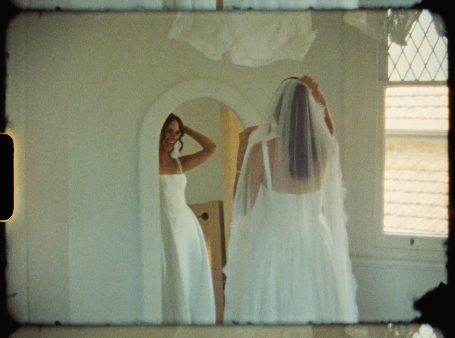 Bride in white gown and veil adjusts her look in a vintage round mirror.