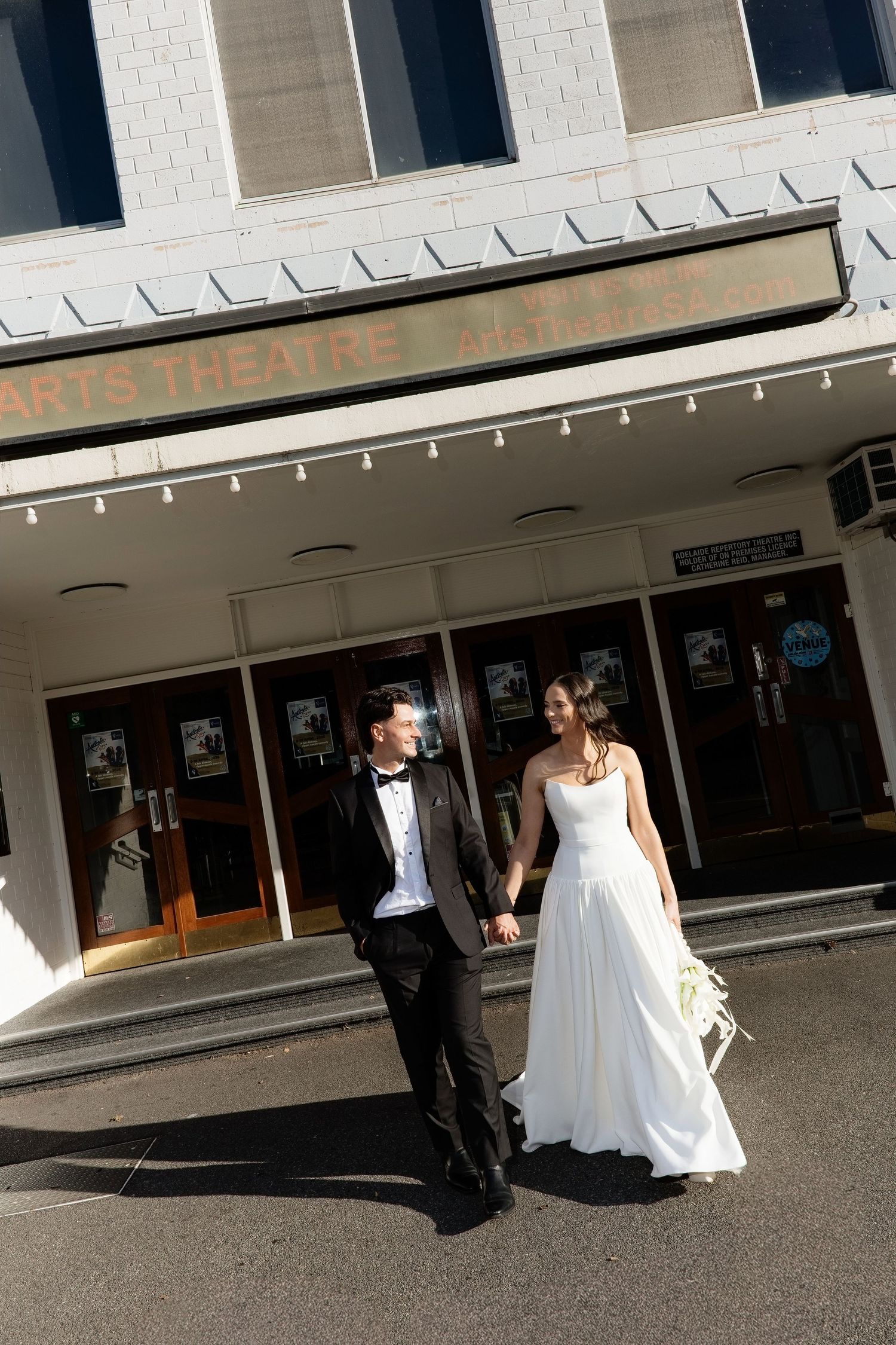 Bride and groom holding hands walking outside Arts Theatre, bride in flowing white gown carrying bouquet.