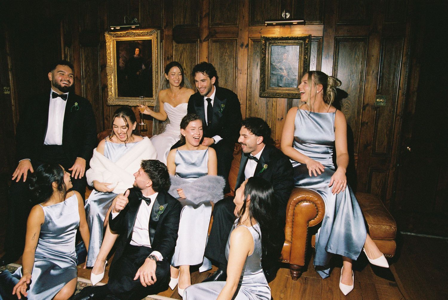 Wedding party in formalwear gathered on leather sofa in wood-paneled room with portrait paintings.