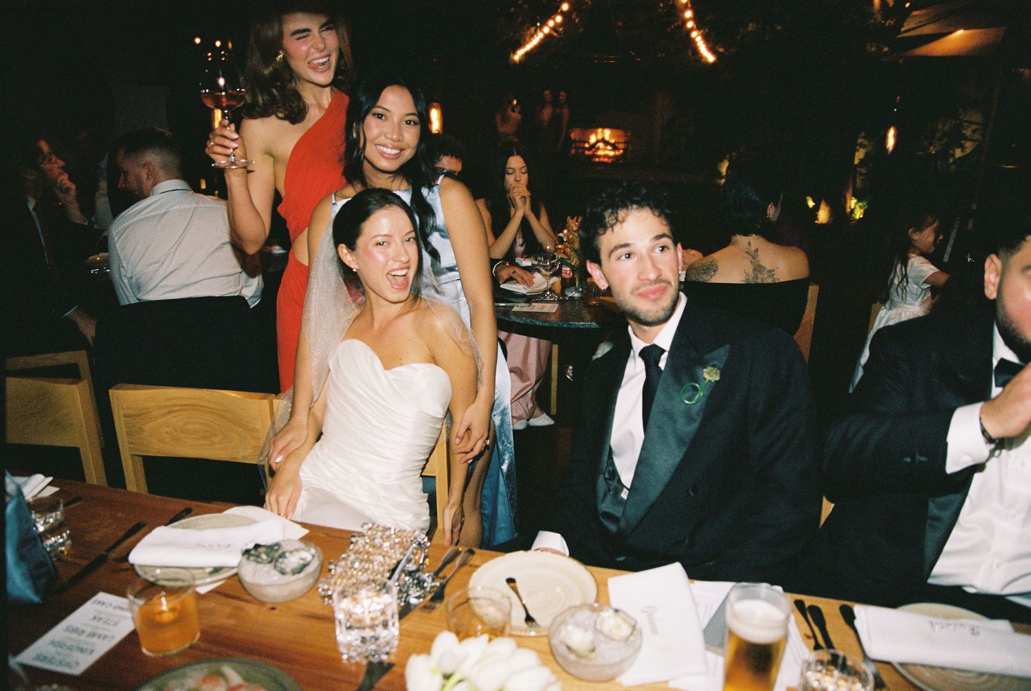 Bride in white strapless gown sits at wedding reception table with groom in tuxedo surrounded by smiling guests.
