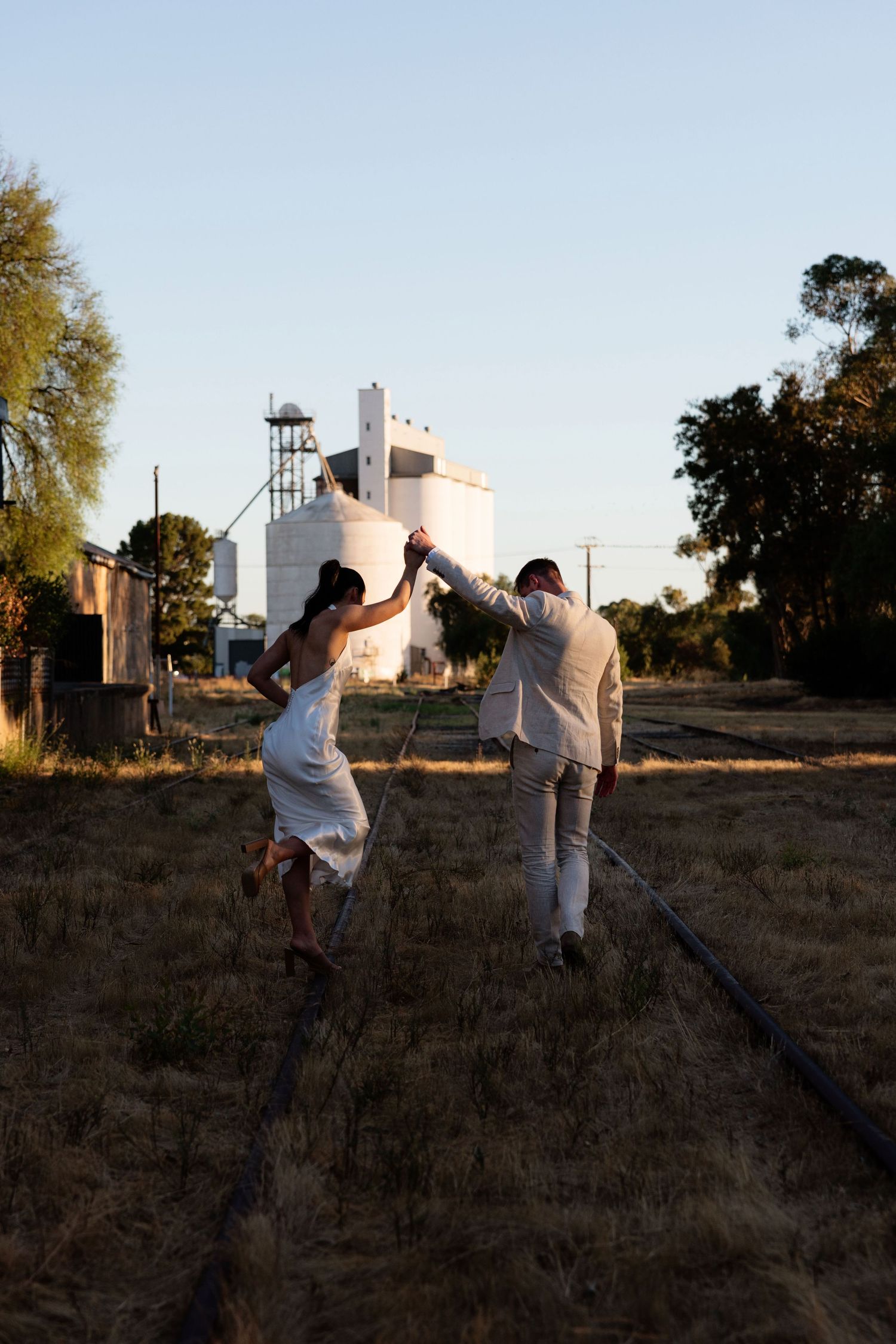 Bride and groom dancing joyfully on rustic railway tracks near grain silos at sunset.