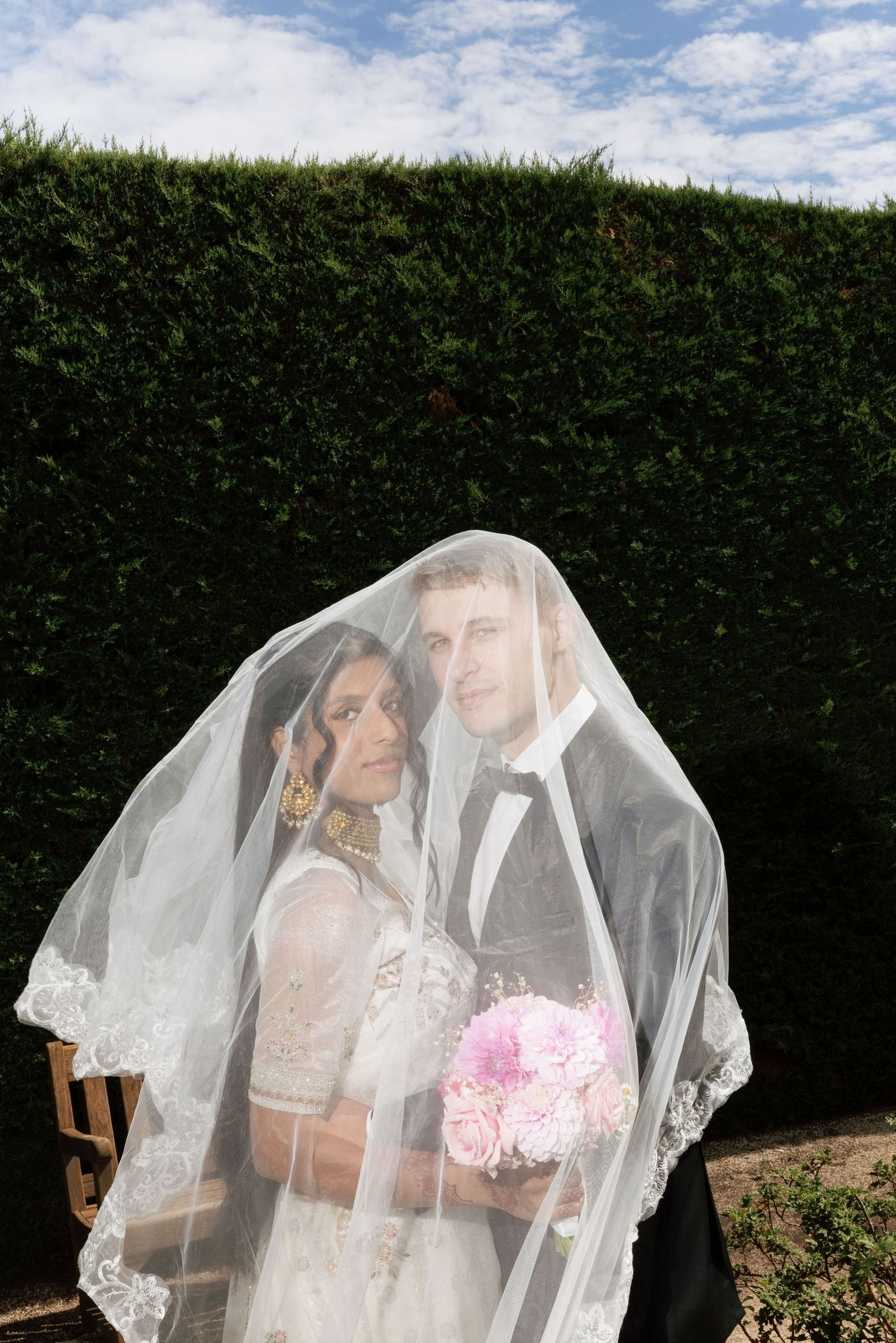 Bride and groom sharing romantic moment under flowing white veil, holding pink bouquet, outdoor garden setting.