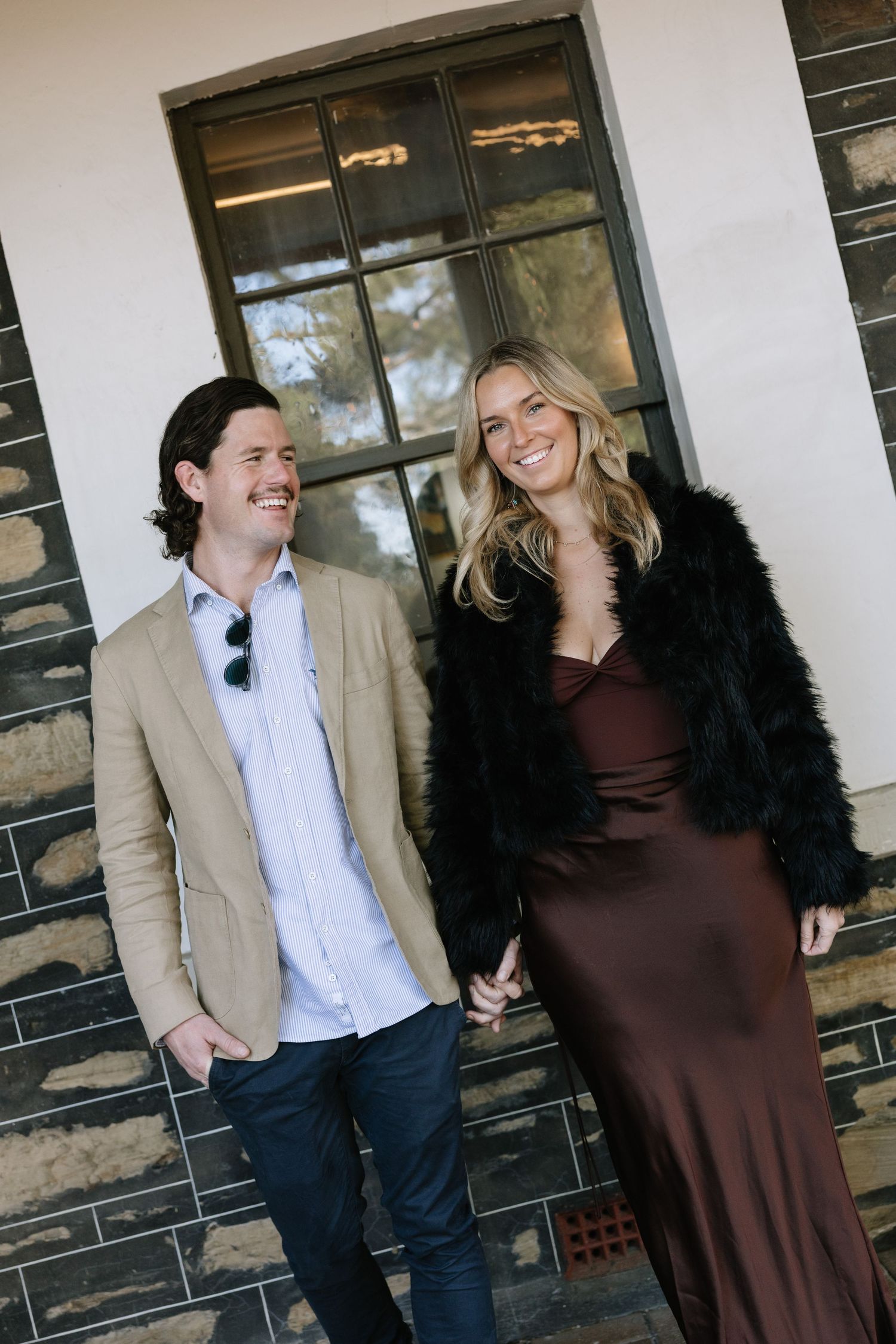 Smiling couple holding hands outdoors, man in tan blazer and woman in brown satin dress with black fur coat.
