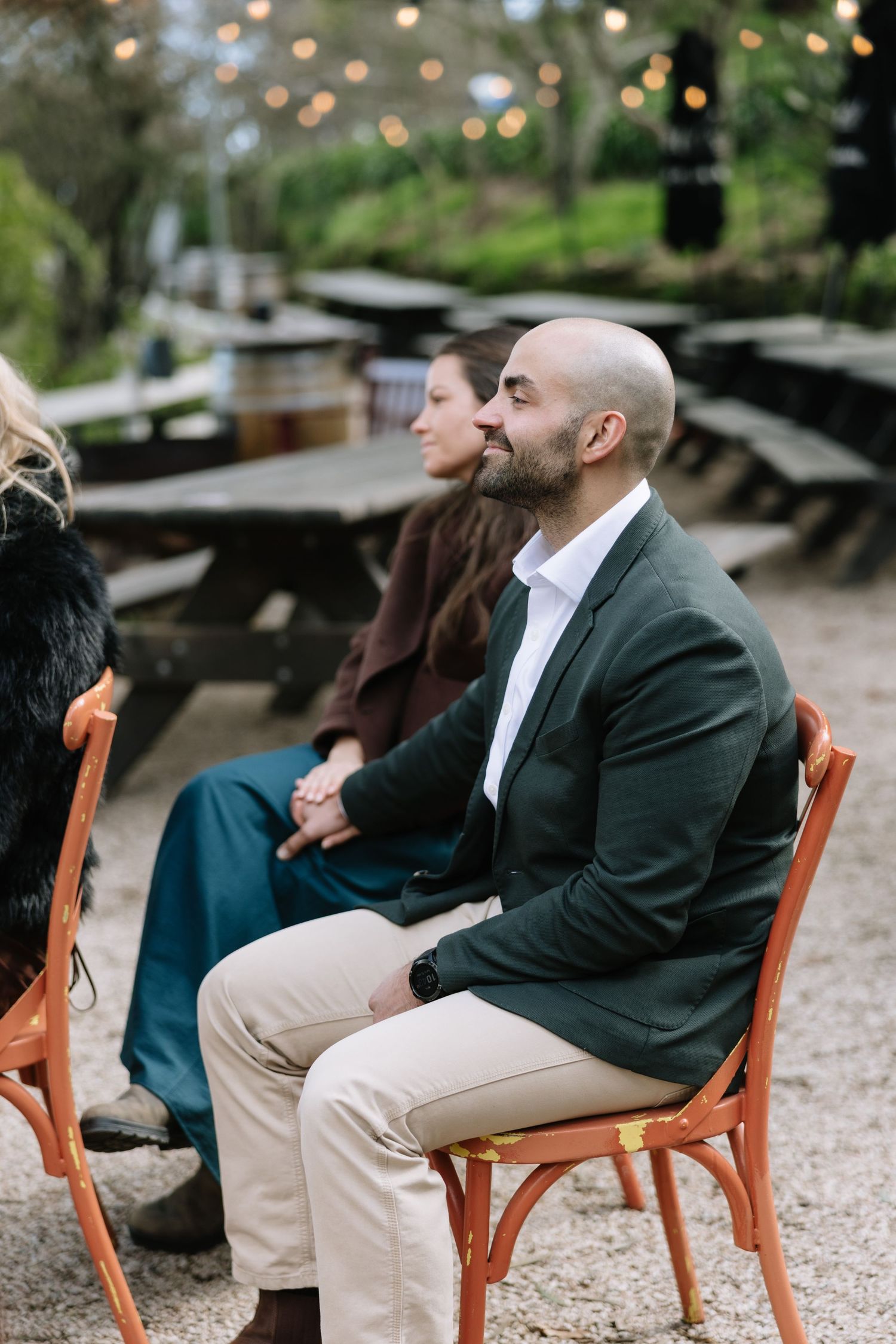 A bald bearded man in a dark blazer sits attentively on an orange chair at an outdoor event.