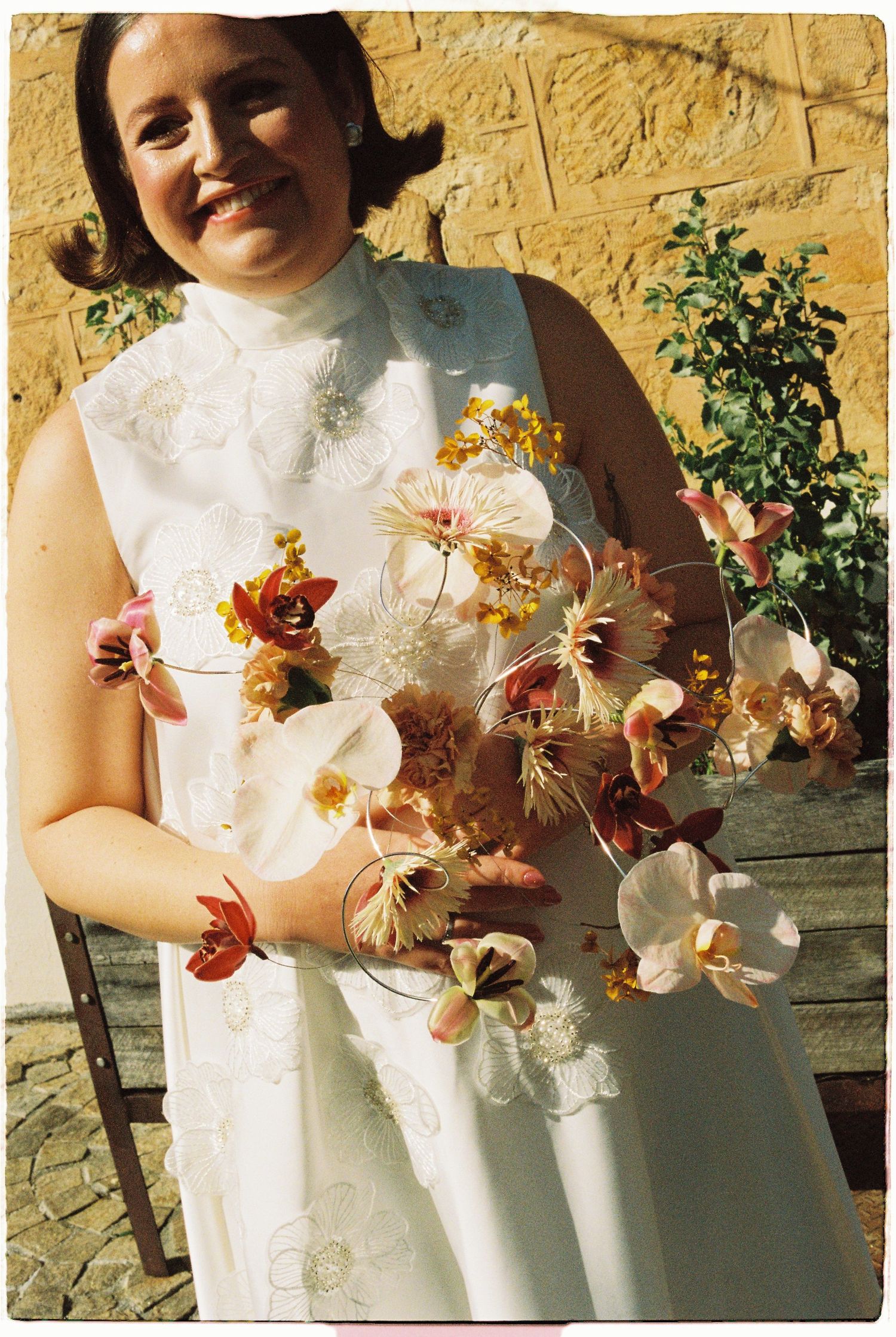 Smiling bride in white embroidered dress holding a cascading bouquet of colorful orchids outdoors.