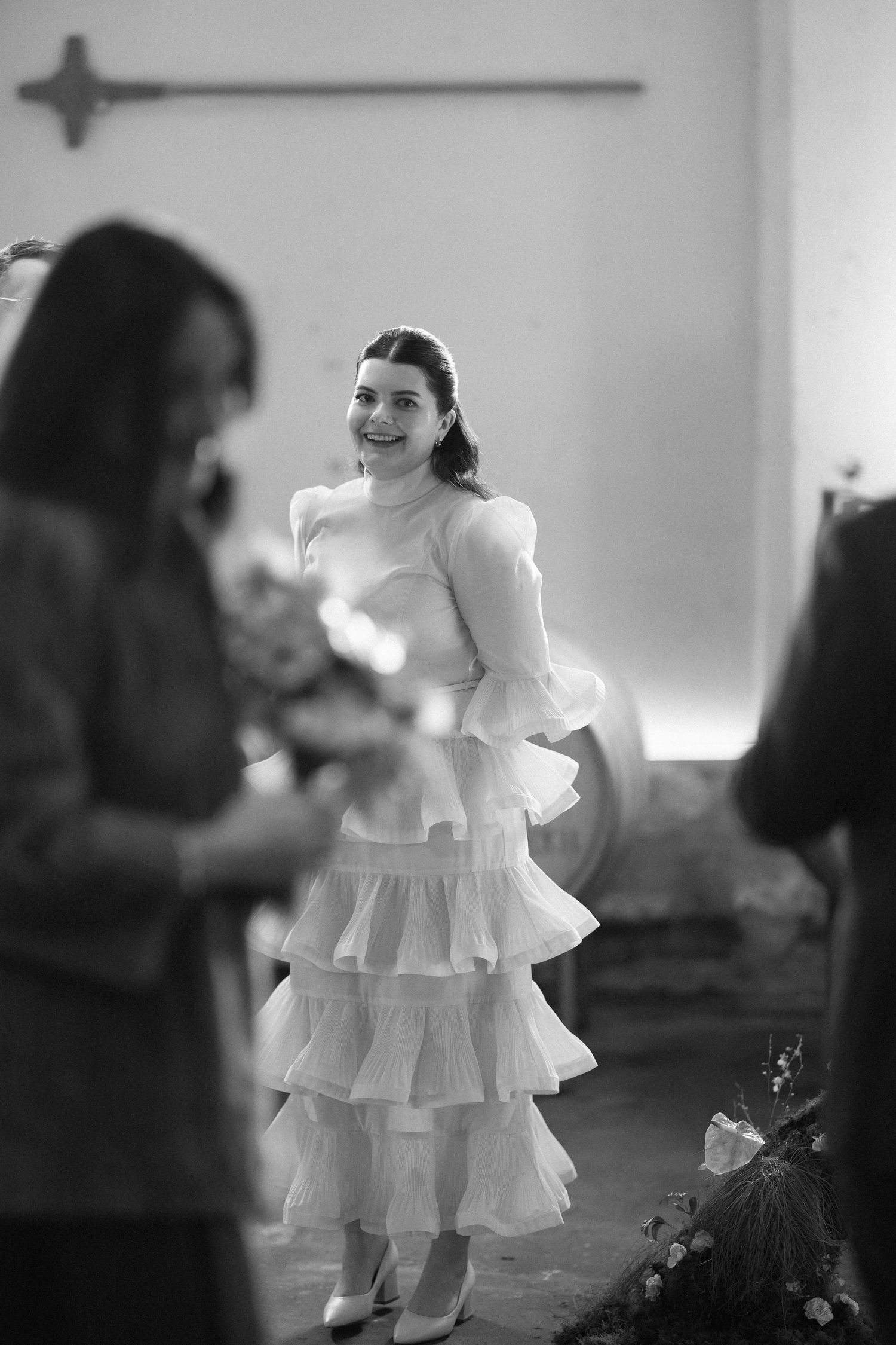 Smiling bride in tiered ruffled white gown standing in winery ceremony space in black and white.