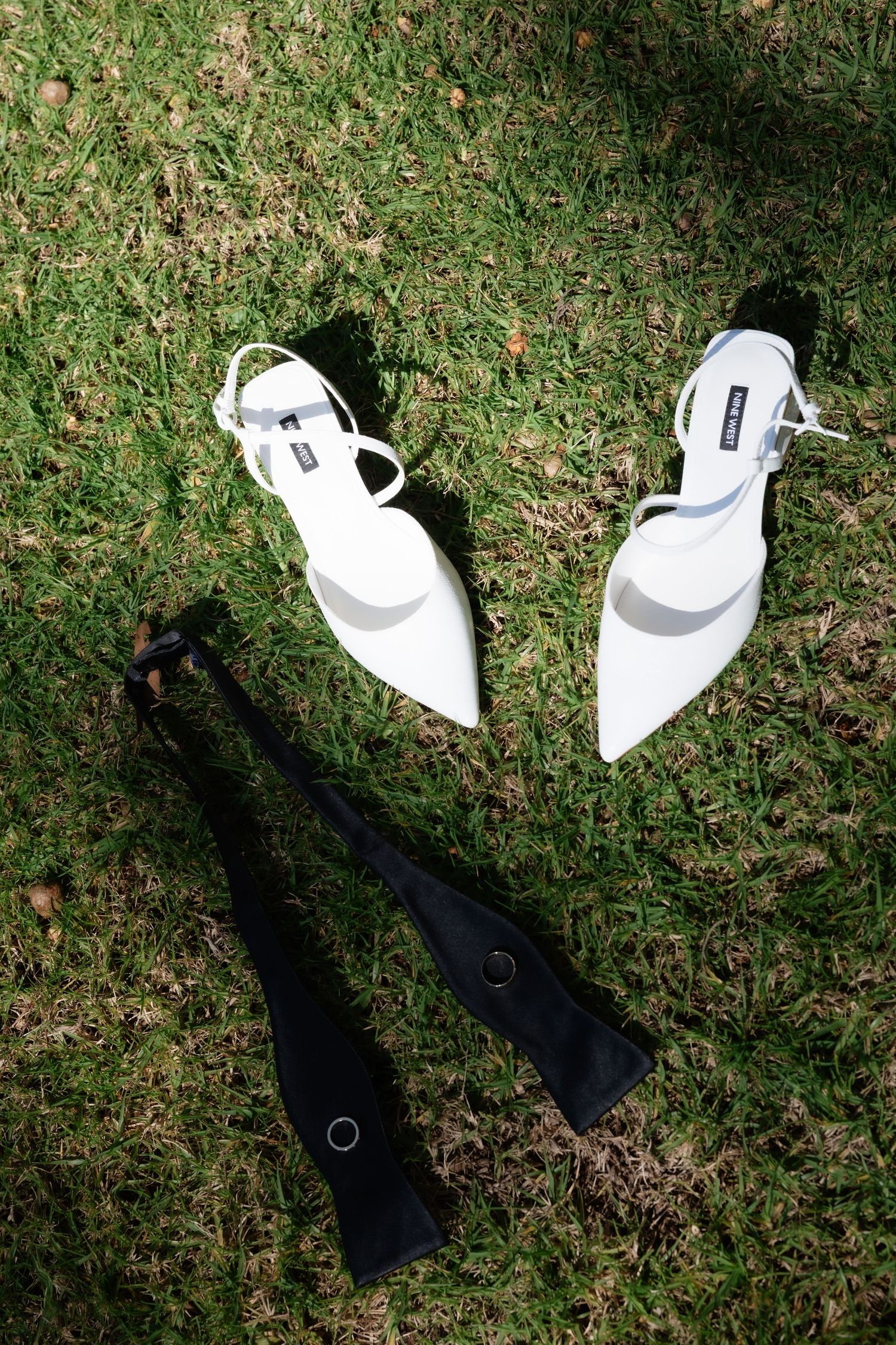 White bridal heels and black bow tie with wedding ring on green grass.