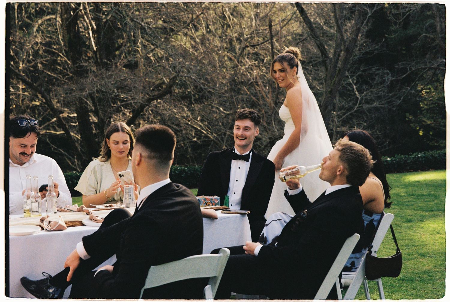 Wedding guests in formal attire laugh and converse at outdoor reception table while bride stands giving speech.
