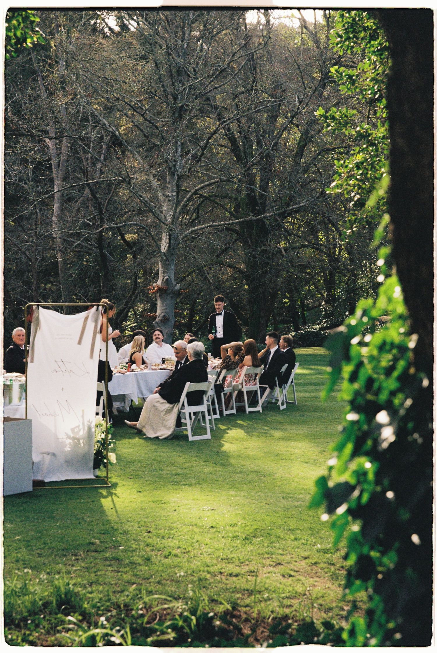 Elegant outdoor wedding reception set on green lawn surrounded by tall trees.