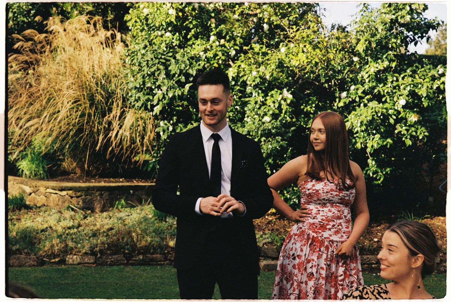 Elegant wedding guest in black suit stands with woman in floral dress in garden setting.