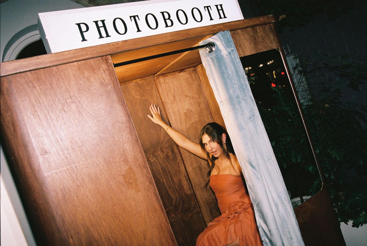 Woman in orange dress posing at vintage wooden photo booth at evening wedding reception.