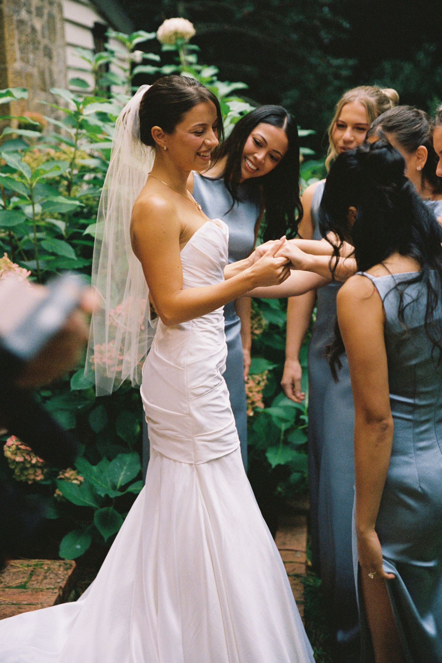 Smiling bride in strapless white gown and veil shares a joyful moment with bridesmaids in blue dresses.