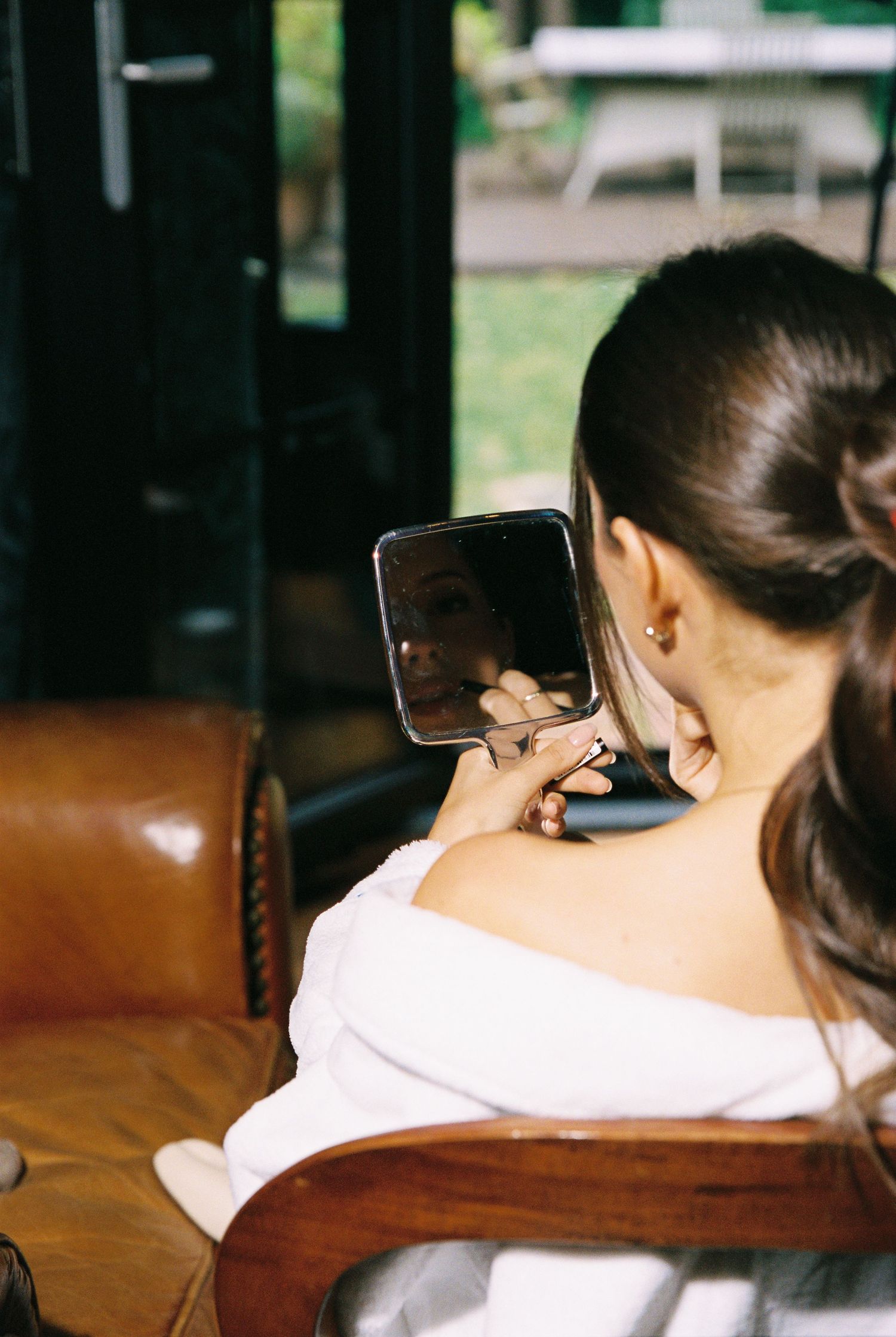 Bride checking makeup in a small mirror while seated on a leather chair.