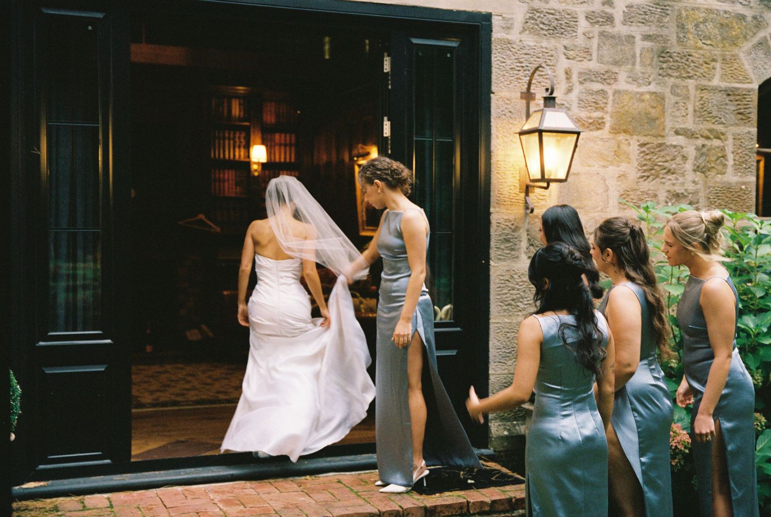 Bride in white gown and veil enters stone building assisted by bridesmaids in blue-gray dresses.