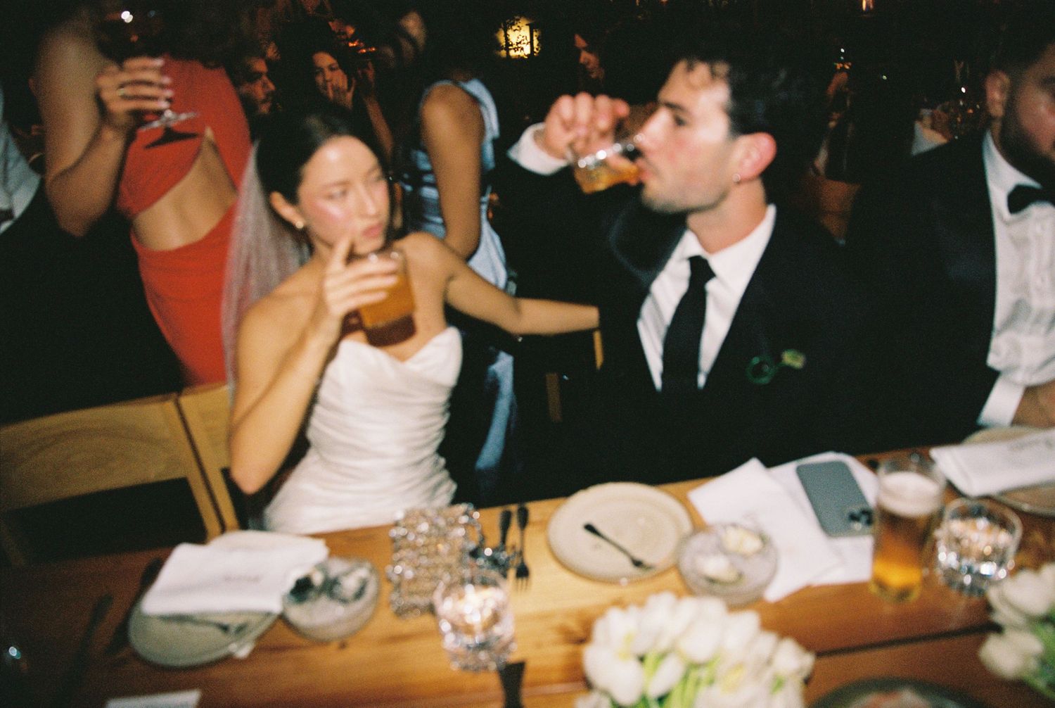 Bride and groom drinking beer together at wedding reception dinner table with flowers.
