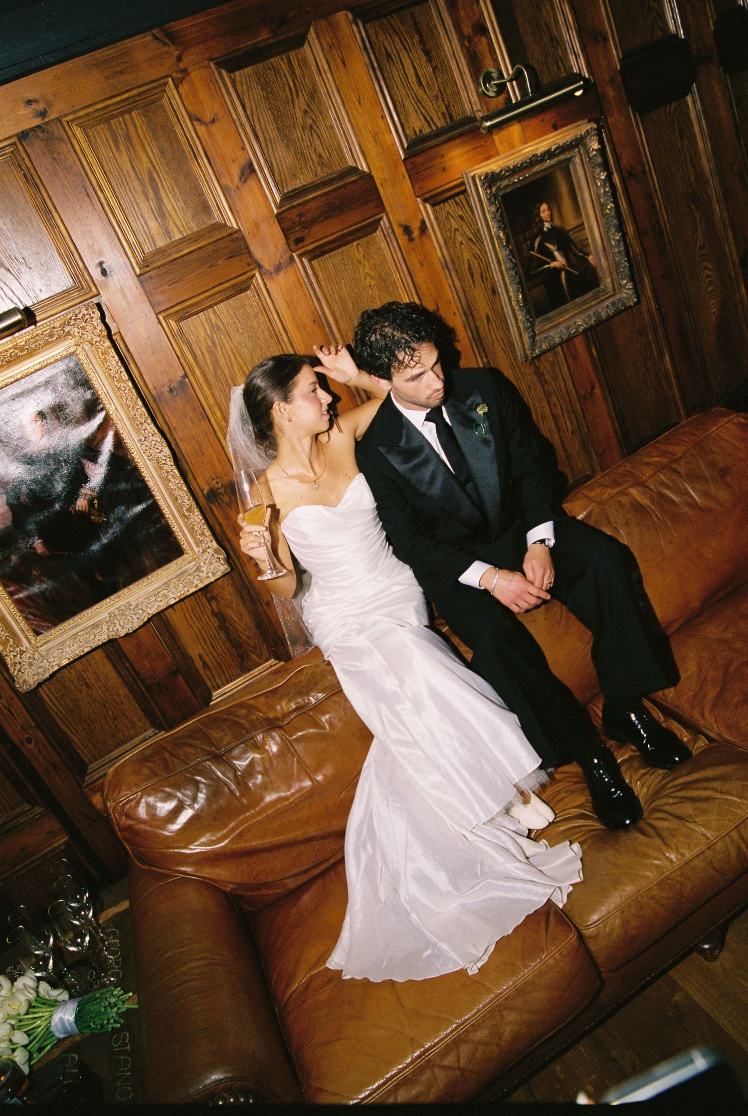 Bride in white gown and groom in tuxedo sharing an intimate moment on a leather couch.