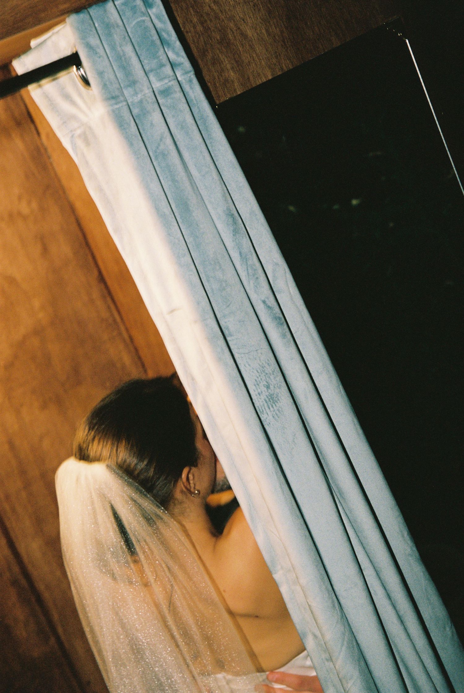 Bride in veil peeks through blue curtain of wedding photobooth.