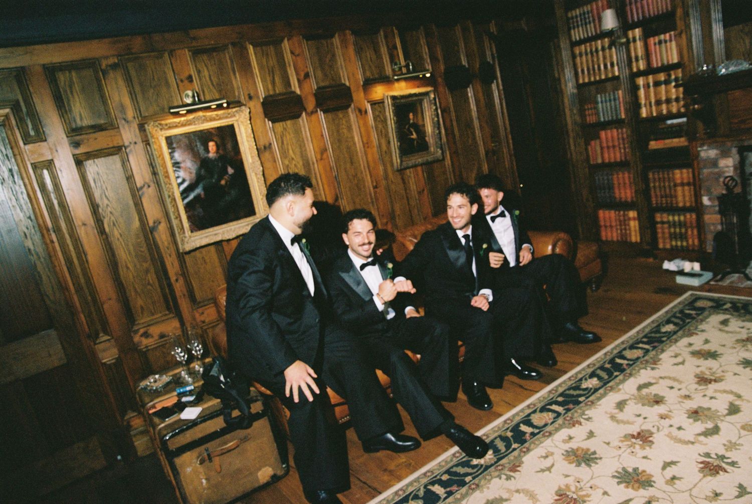 Four groomsmen in black tuxedos laughing together in an elegant wood-paneled library room with bookshelves.