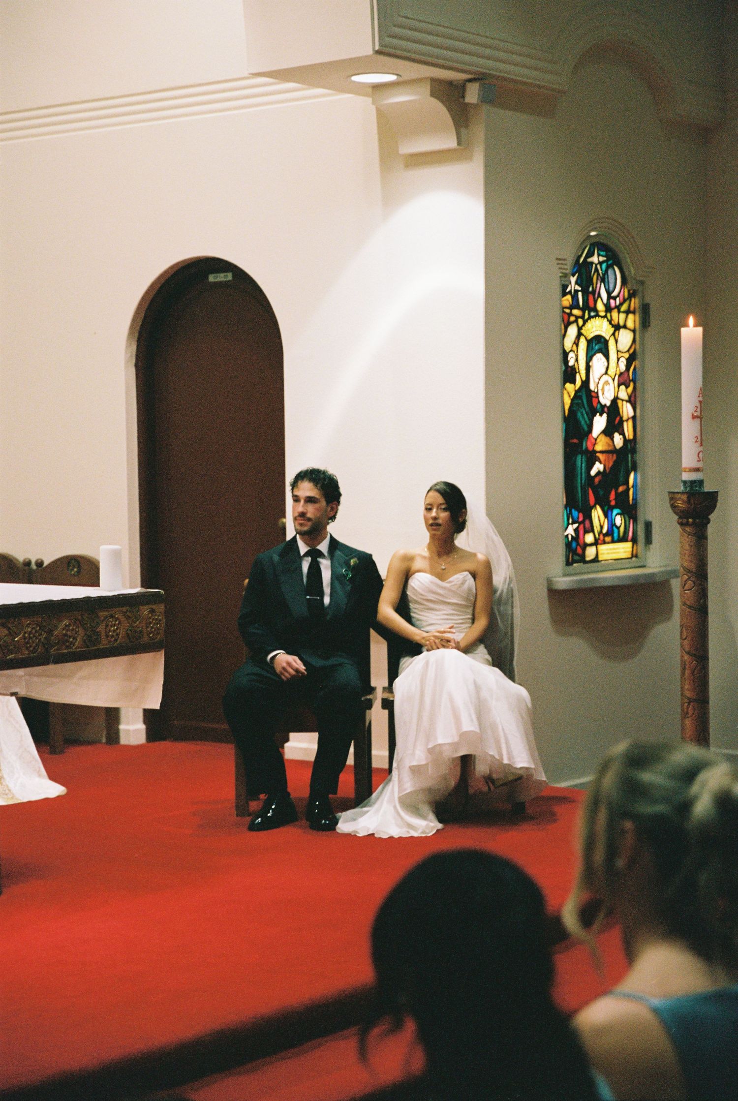 Bride and groom sit together at church altar during wedding ceremony with stained glass window behind.