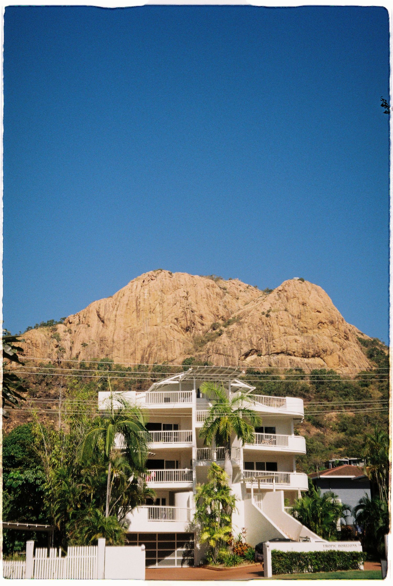 White apartment building with balconies beneath a large rocky mountain peak against a clear blue sky.