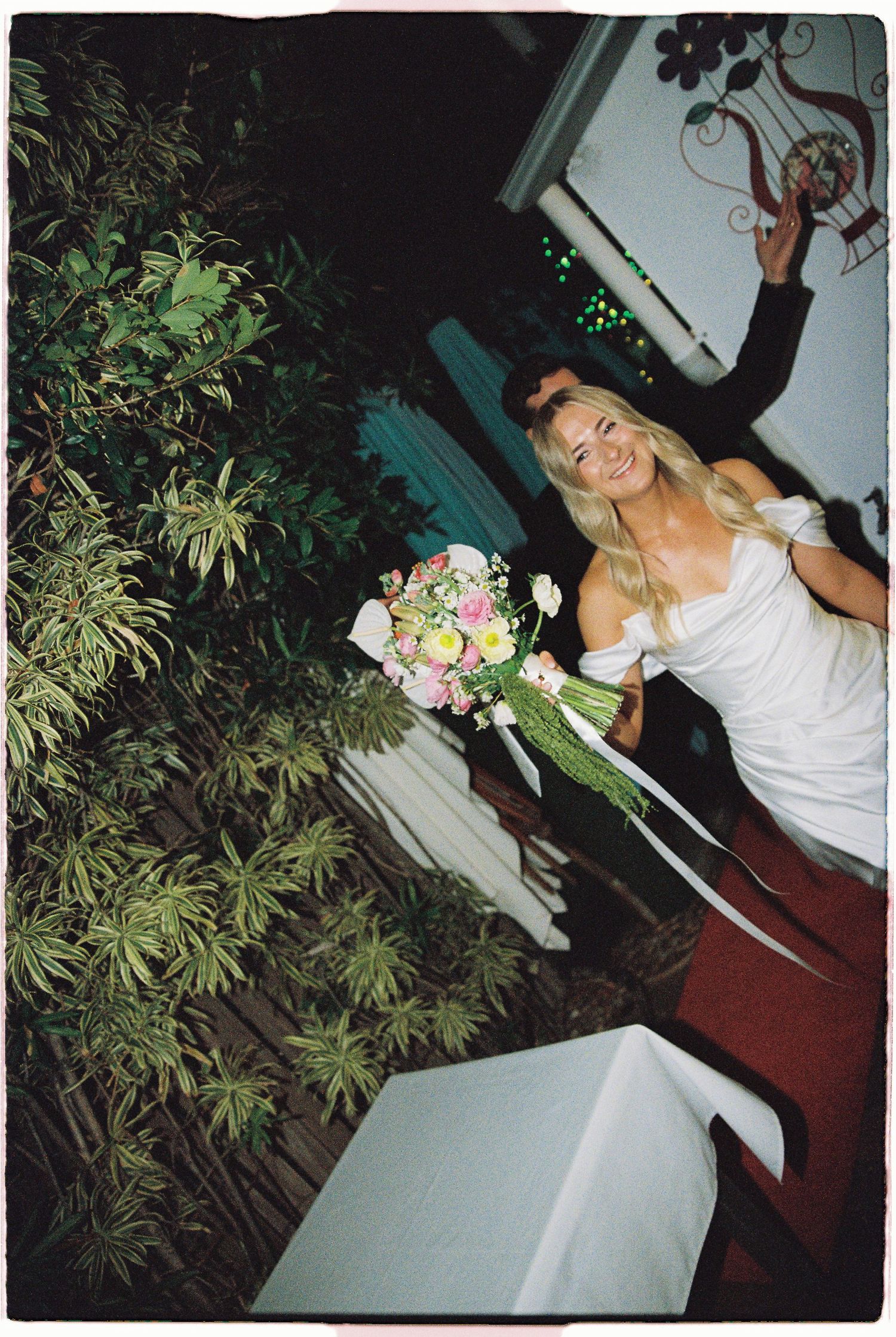 Smiling bride in off-shoulder white gown holds colorful bouquet at outdoor wedding venue.