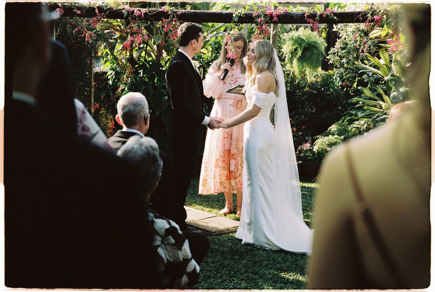 Bride and groom exchange vows outdoors under floral arch with officiant in garden wedding ceremony.