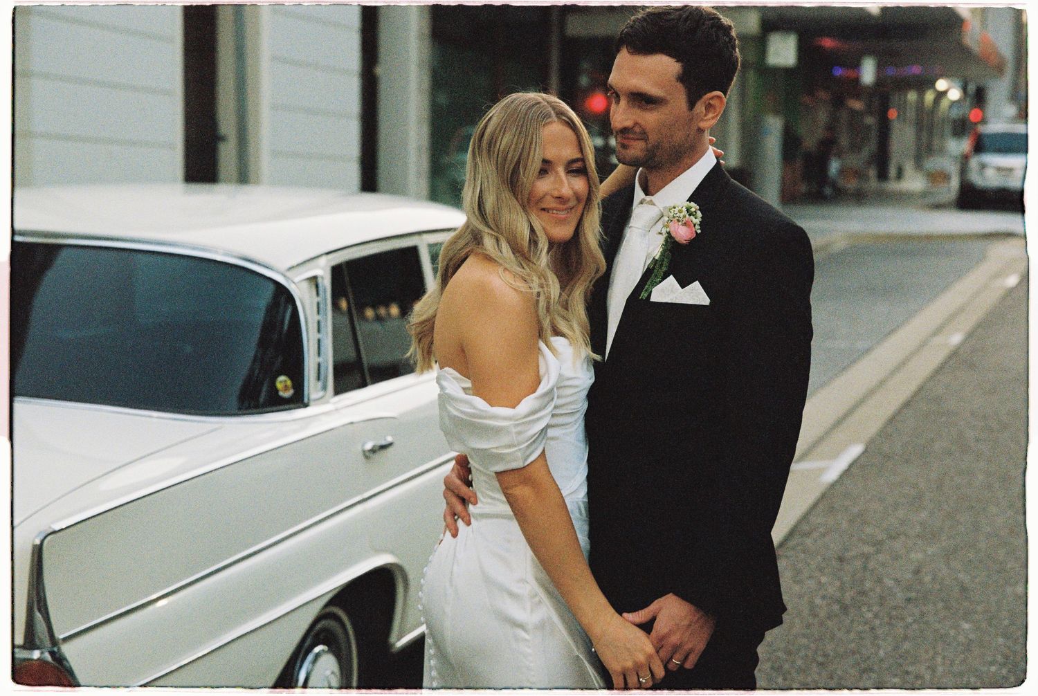 Bride and groom posing beside a classic white vintage car on a city street in wedding attire.