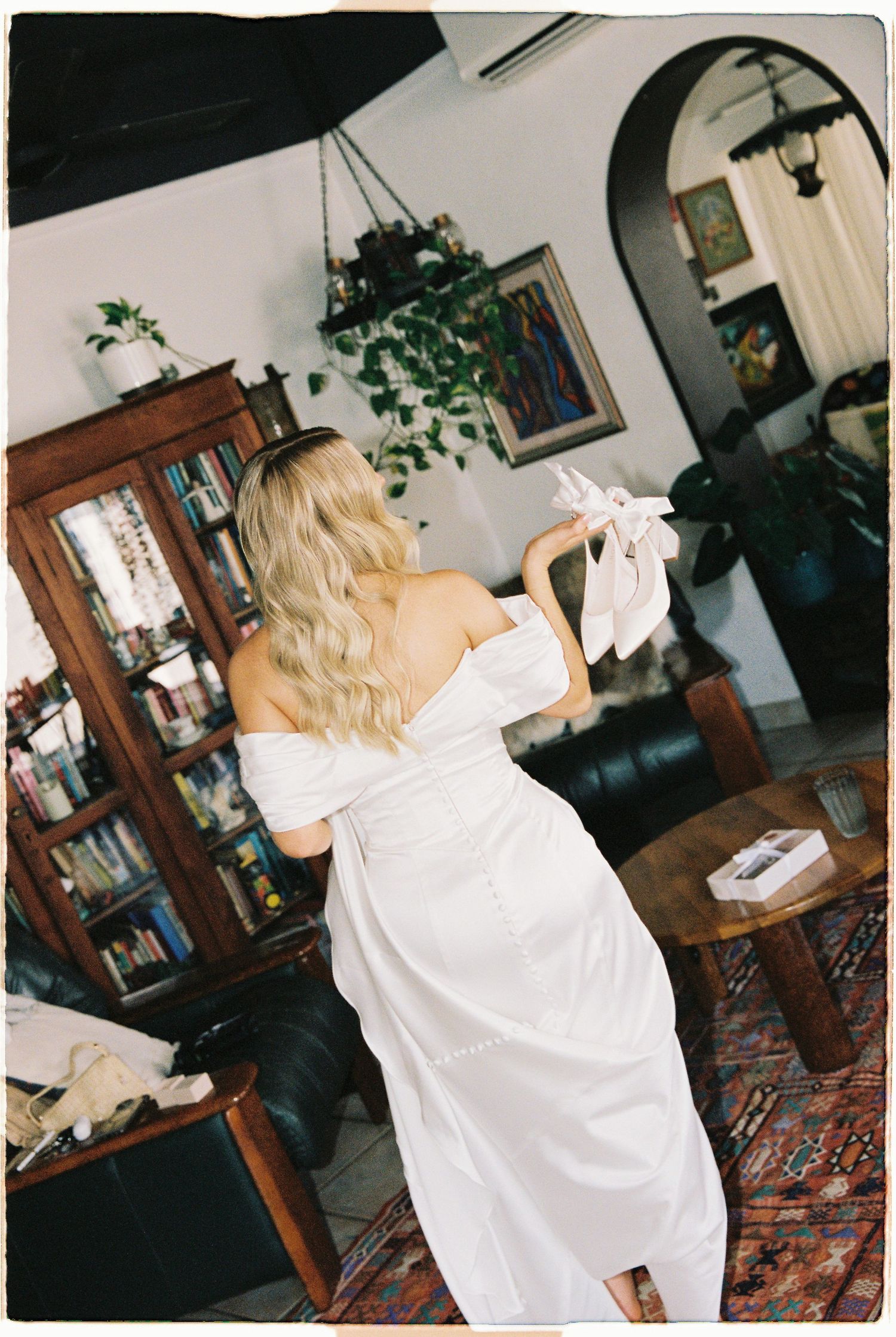 Blonde bride in white off-shoulder gown adjusting her bow detail in a decorated living room.