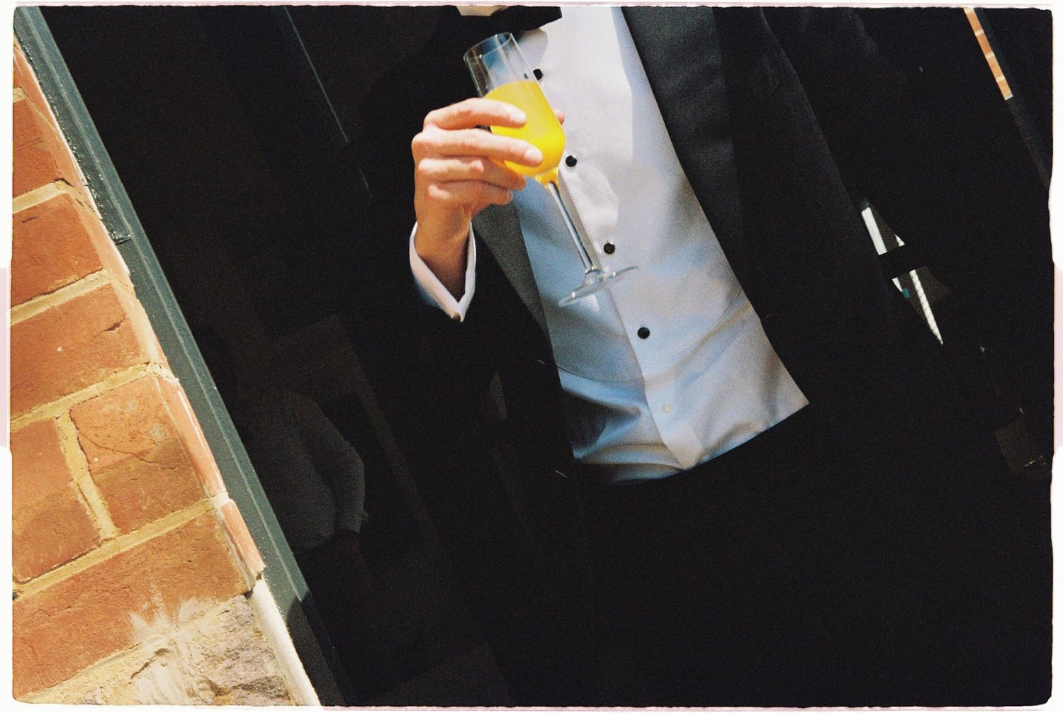 Groom in black tuxedo holding a yellow citrus drink outside brick building at wedding reception.