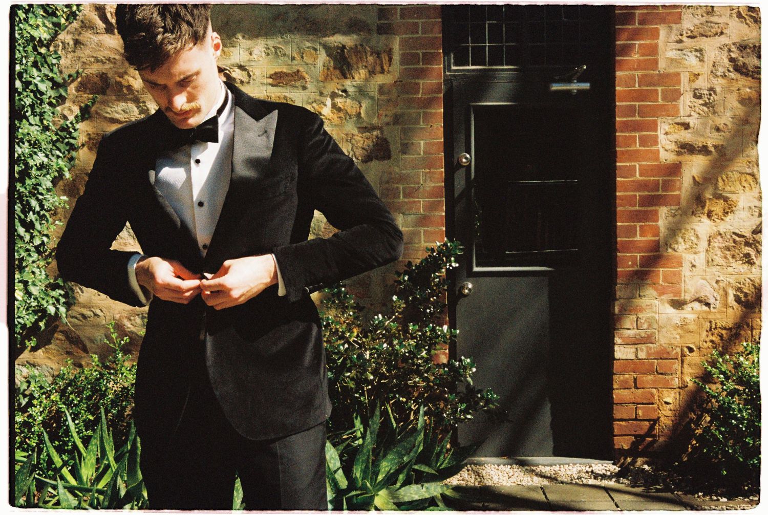 Groom in black tuxedo adjusting jacket buttons outside rustic brick building.