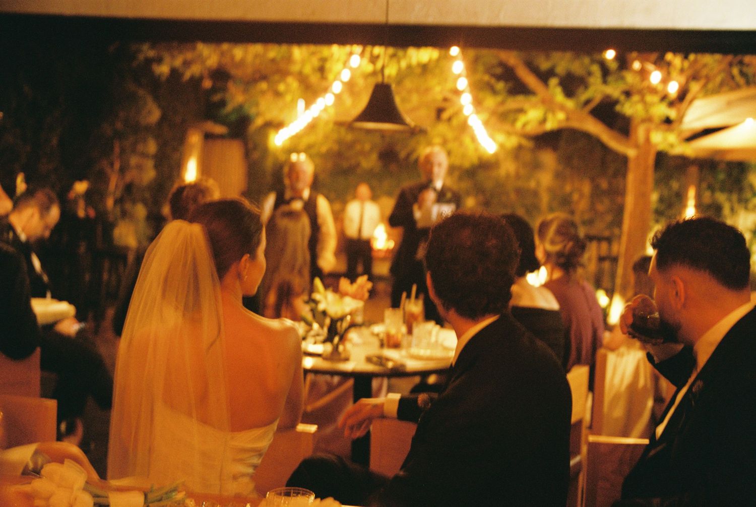 Elegant wedding reception with guests gathered under warm string lights and candles at an outdoor evening venue.
