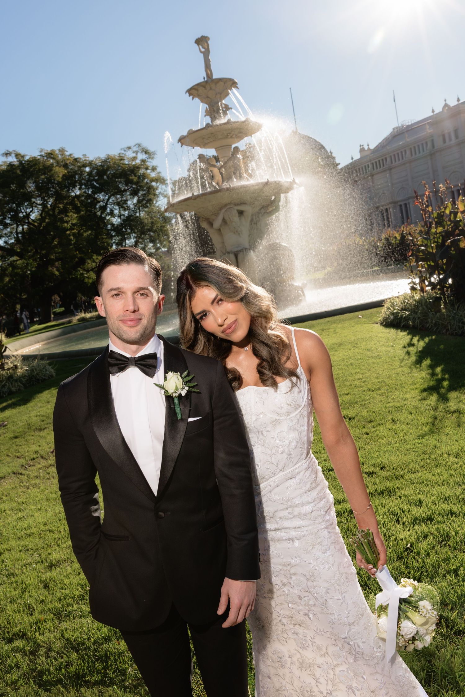 Bride and groom posing in front of ornate fountain in lush garden, groom in black tuxedo, bride in white lace gown.