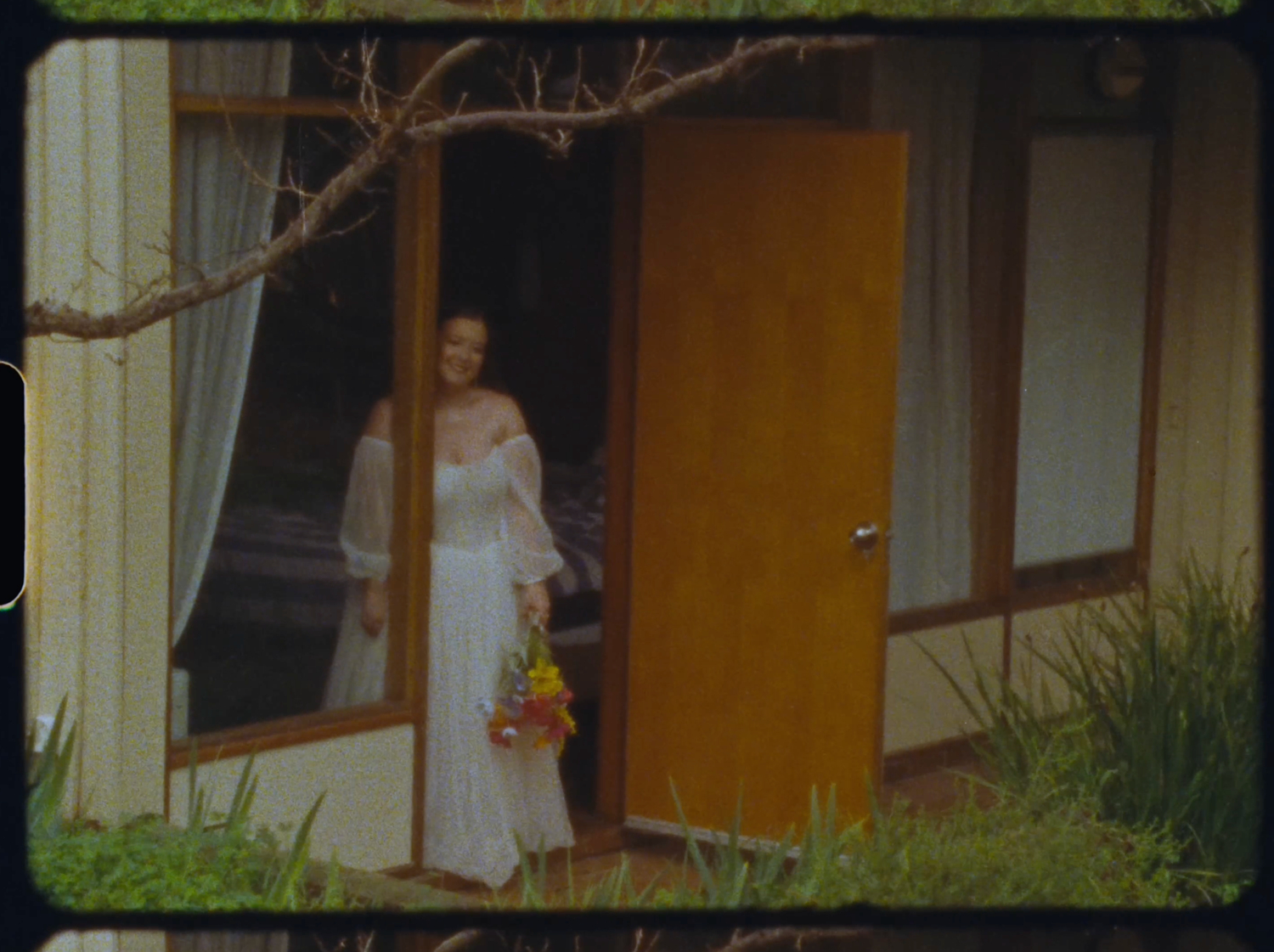 A bride in a white dress holding a bouquet stands in an open wooden doorway of a house.