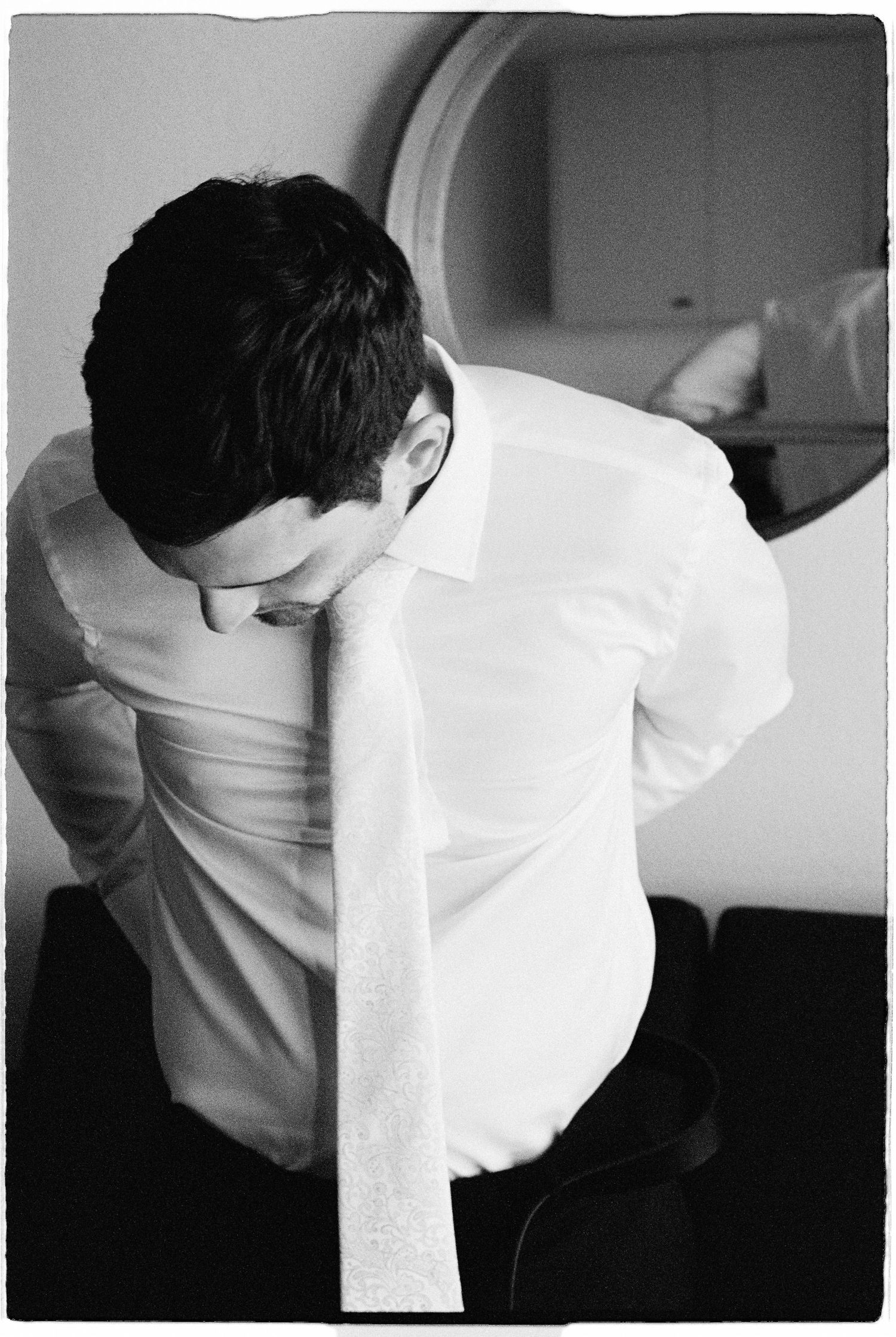 Black and white photo of a groom in a white dress shirt tying his tie while getting ready for his wedding.