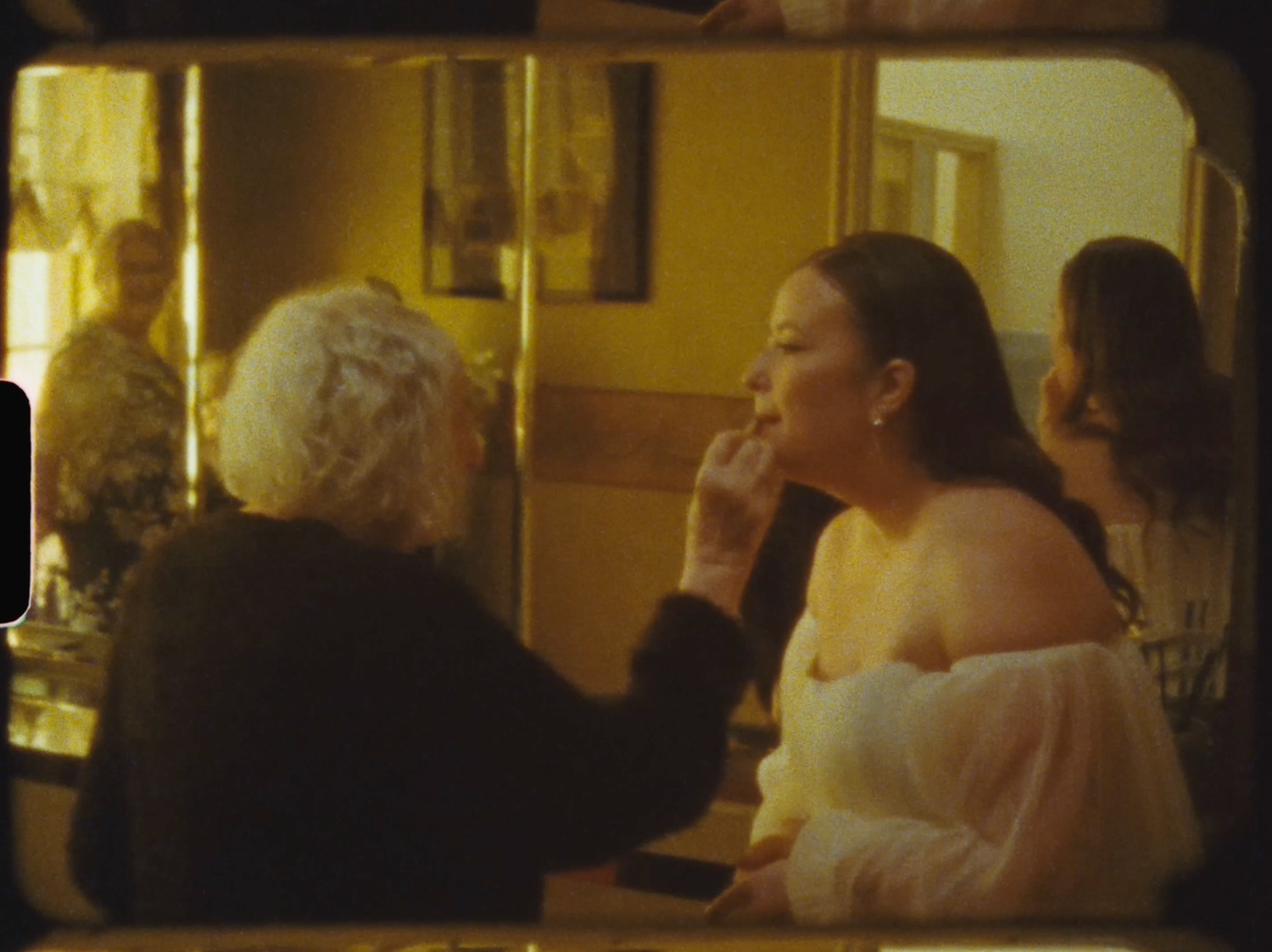 An elderly woman applies makeup to a bride in a warmly lit vintage mirror dressing room.