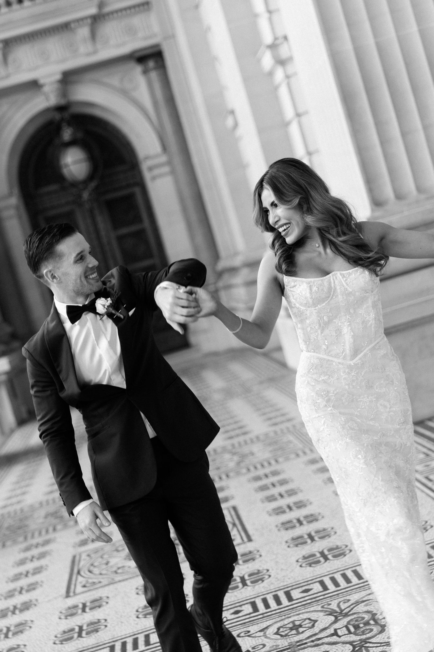 Black and white photo of a laughing bride in a fitted gown and groom in a tuxedo dancing outside a grand building.