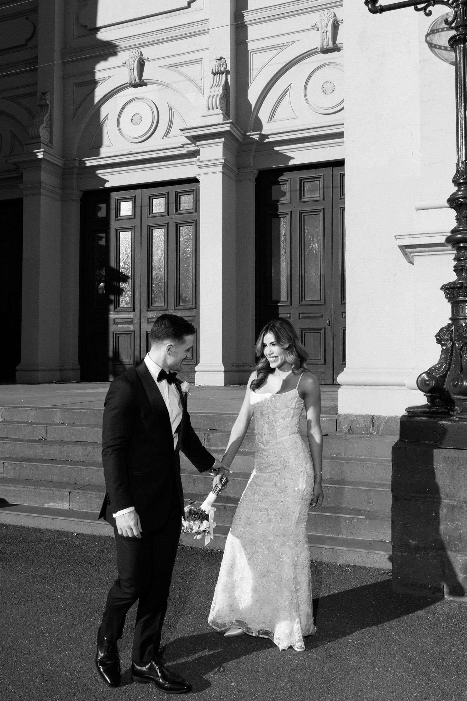 Black and white photo of a bride and groom holding hands outside an elegant building with ornate architecture.