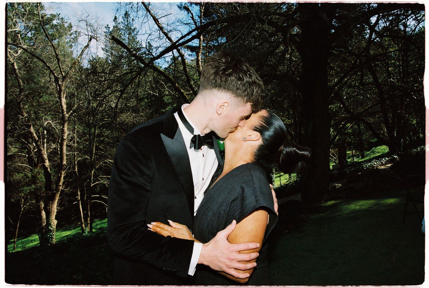 Bride and groom sharing a romantic kiss outdoors surrounded by lush green trees.