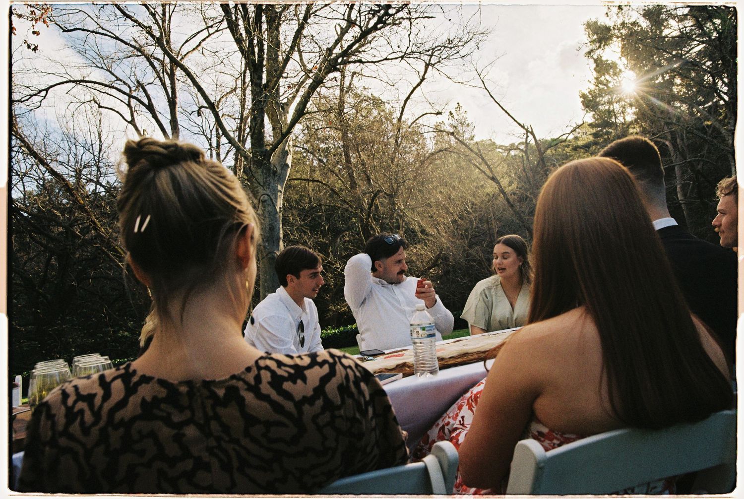 Wedding guests gathered around an outdoor dining table in a sunlit garden setting.