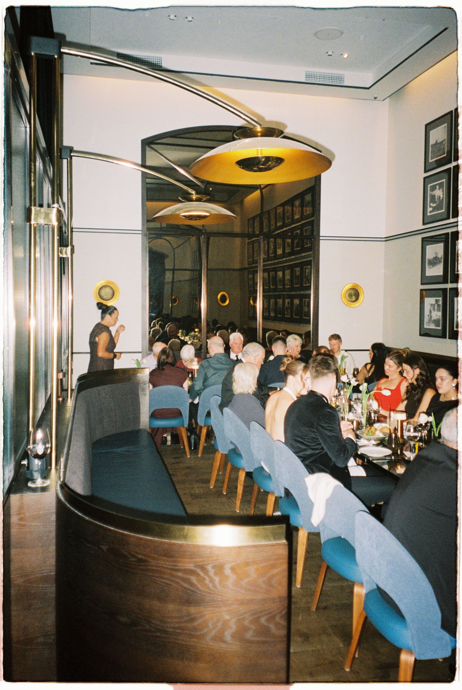 Elegant restaurant dining room filled with wedding guests seated at long table under warm pendant lights.