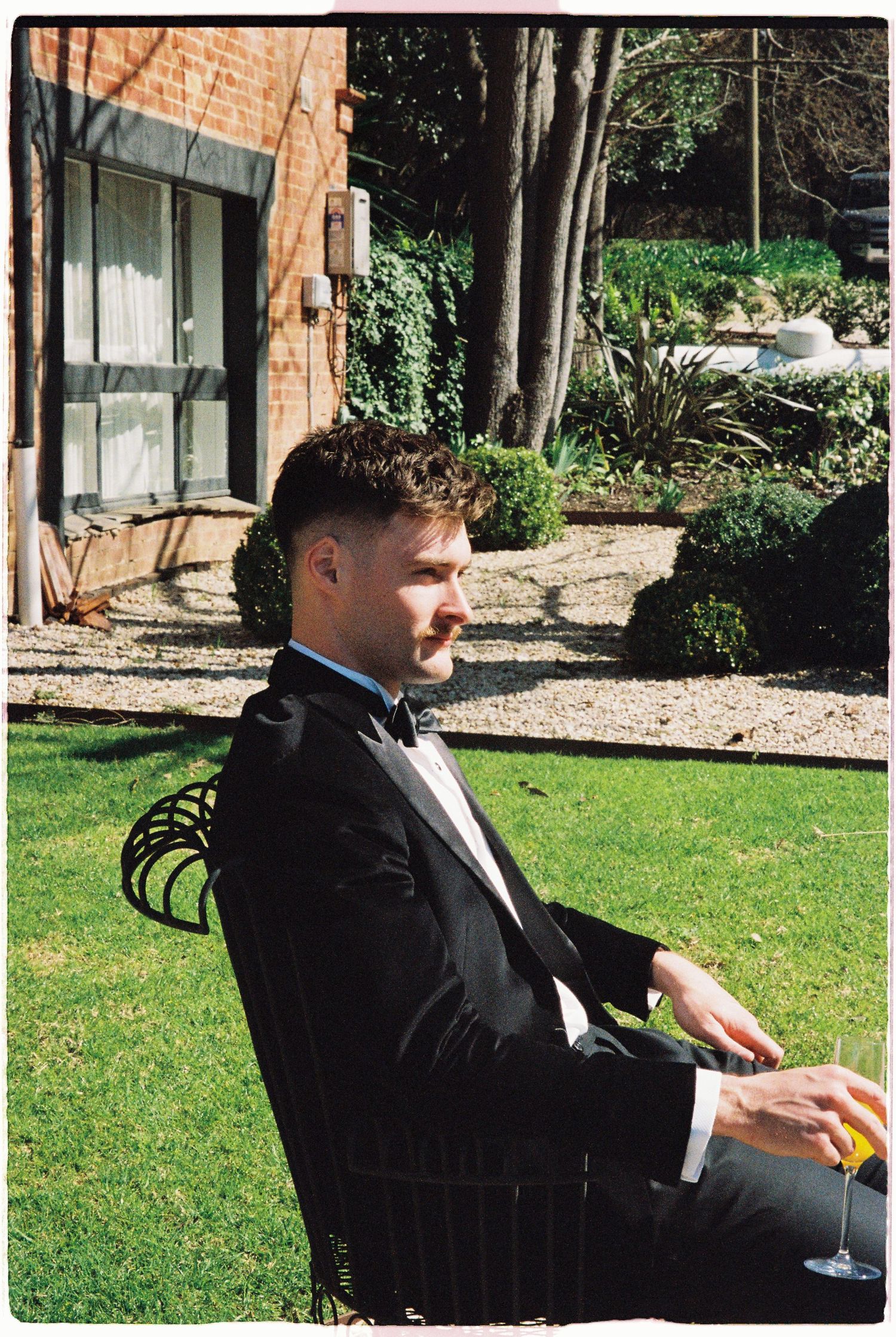 Groom in black tuxedo relaxing in garden chair holding champagne glass outside brick wedding venue.