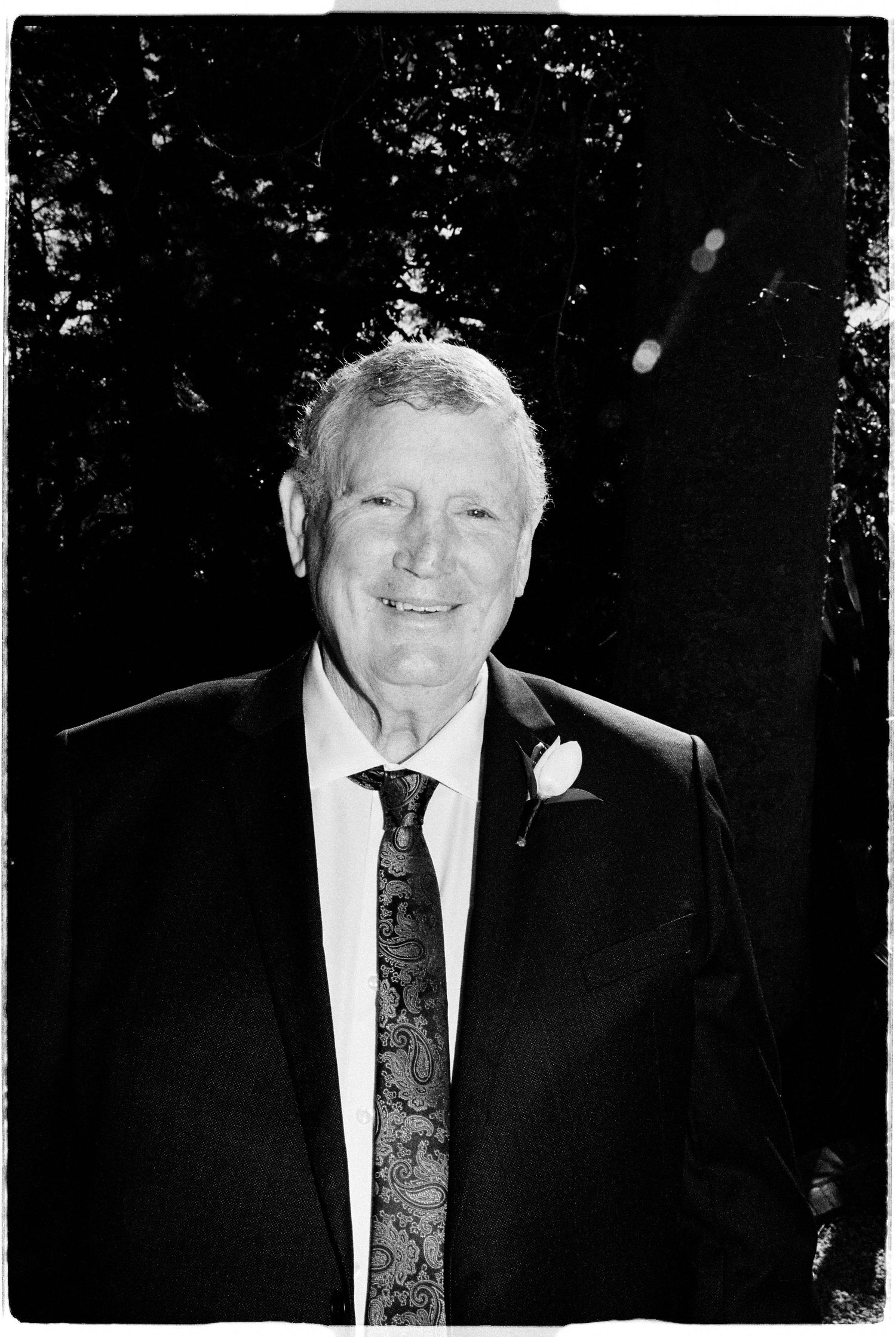 Black and white portrait of a smiling older man in a suit with a floral tie and boutonniere outdoors.