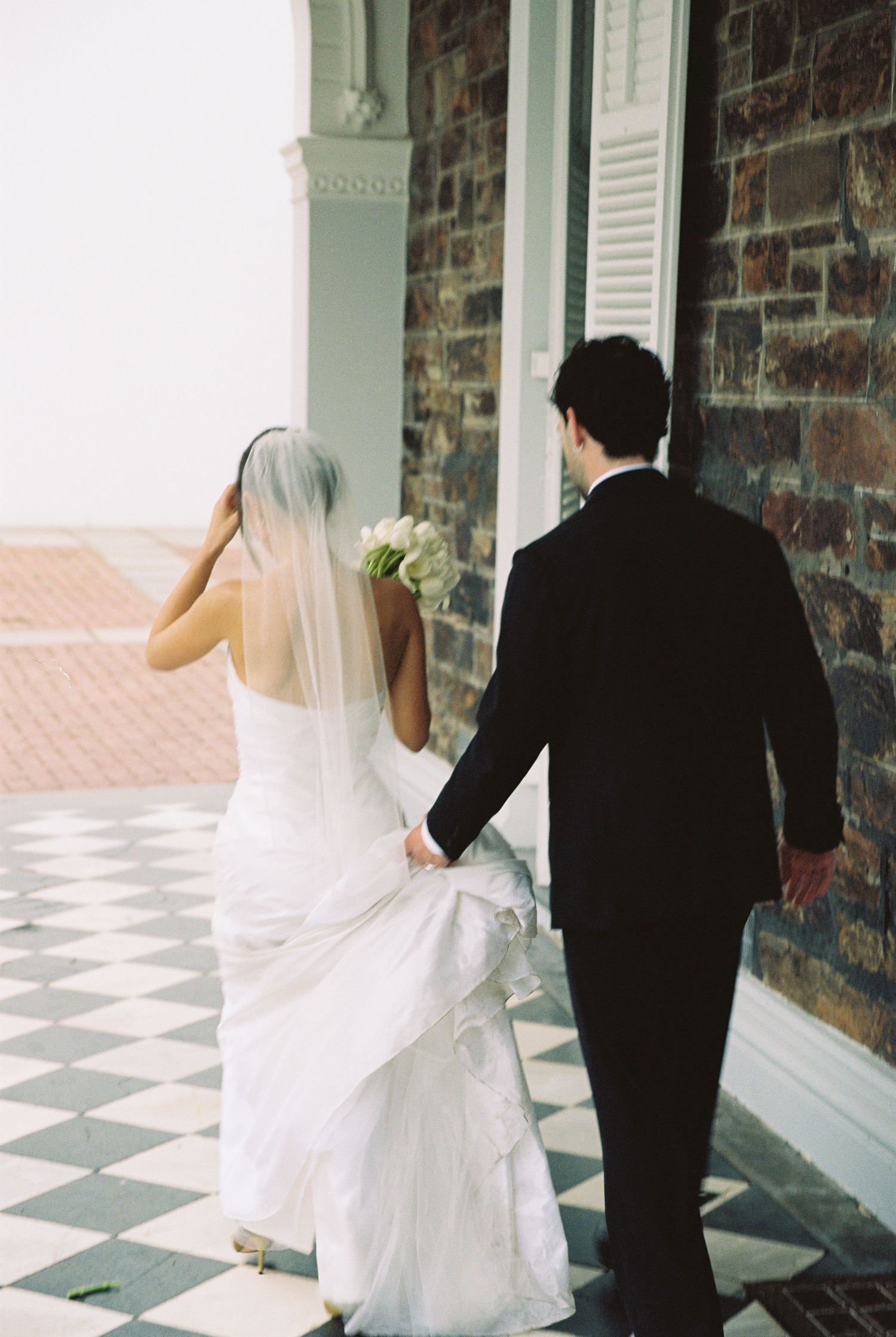 Bride and groom walking hand in hand on checkered floor, bride in white gown with veil holding bouquet.