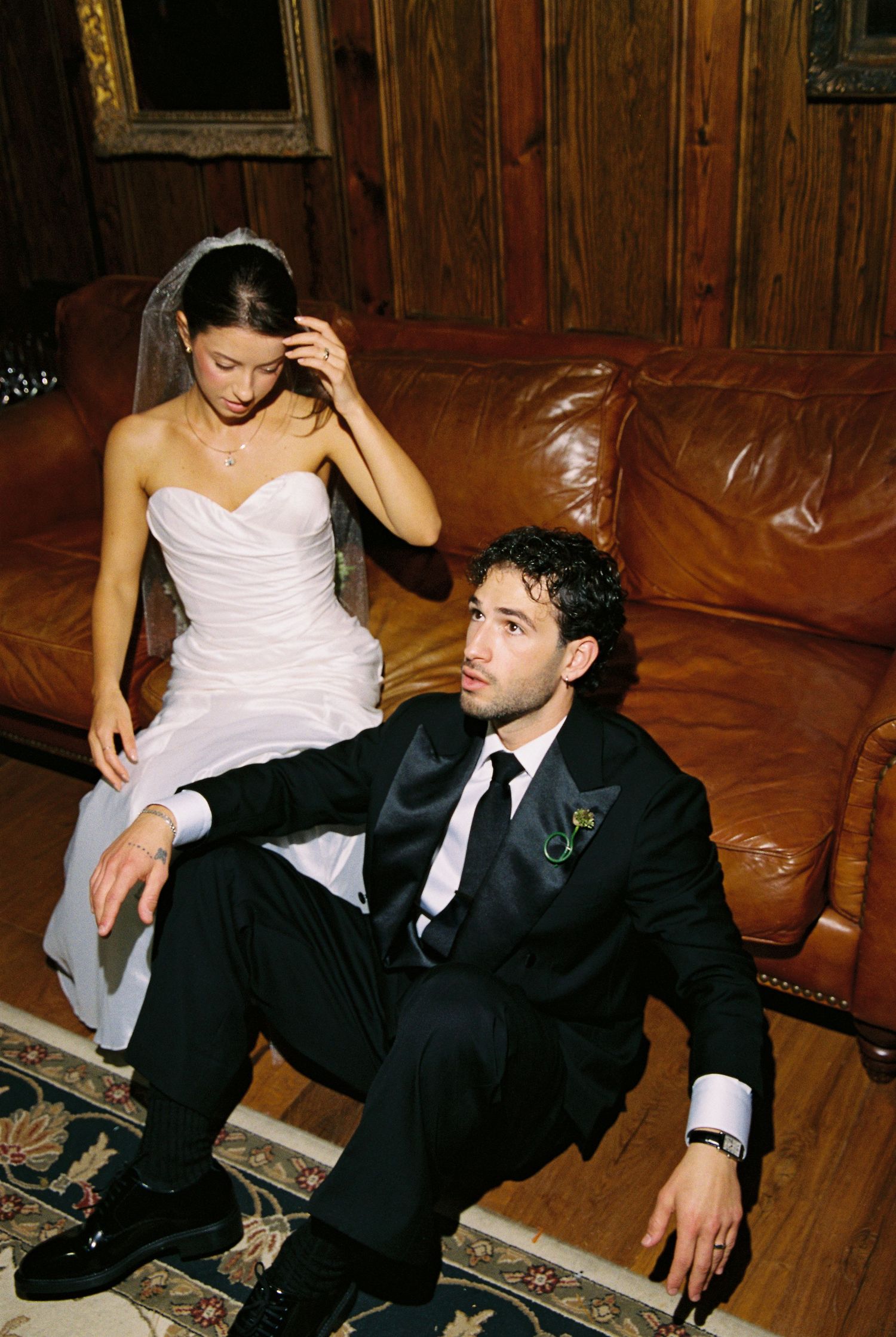 Bride and groom posing on leather couch in rustic wood-paneled room.