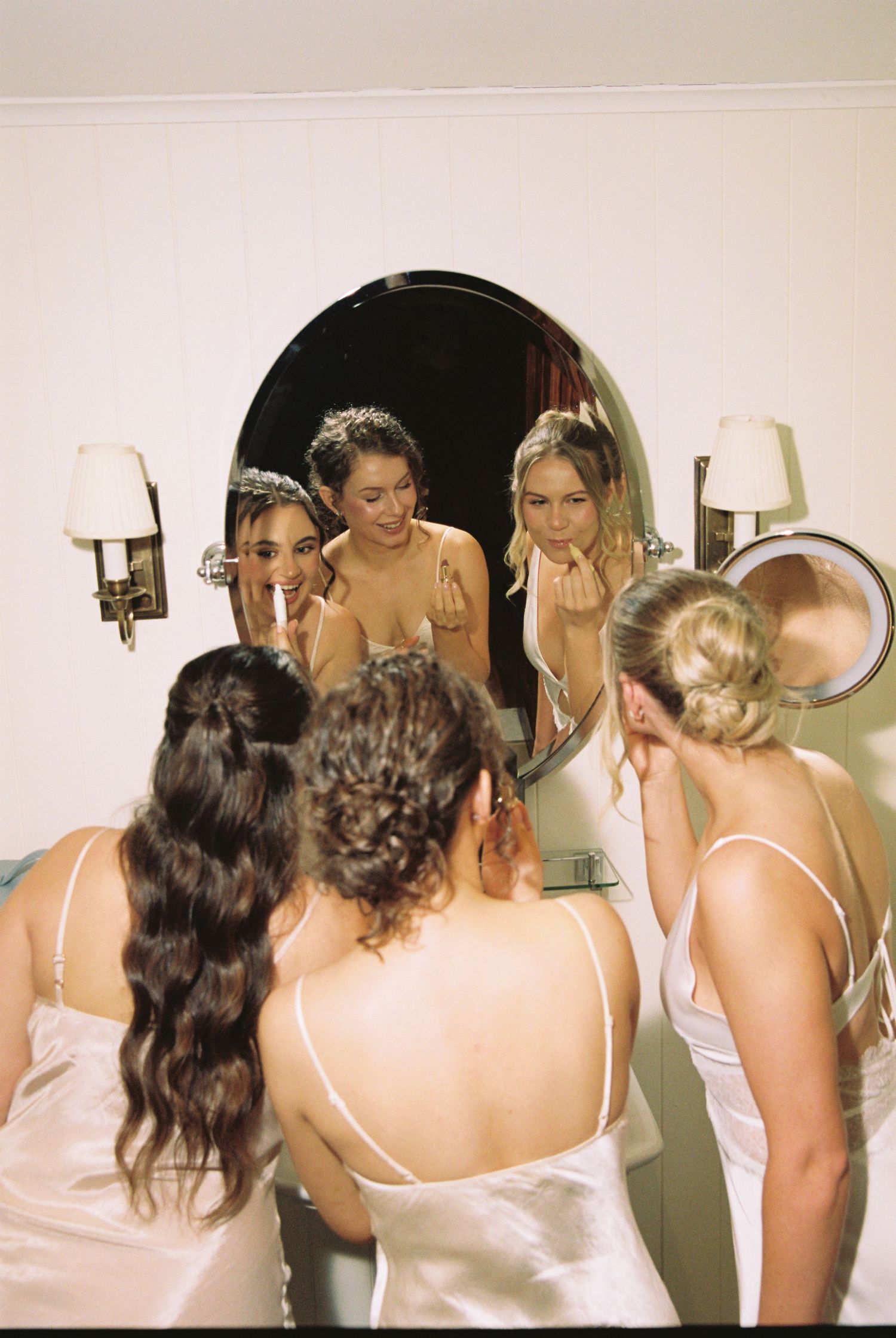 Bridesmaids in white slip dresses gathered around a round mirror doing makeup together.